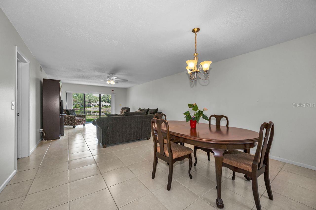 Dining and living room with tiled floors, wooden dining table, dark sofas, and water visible through sliding glass doors.