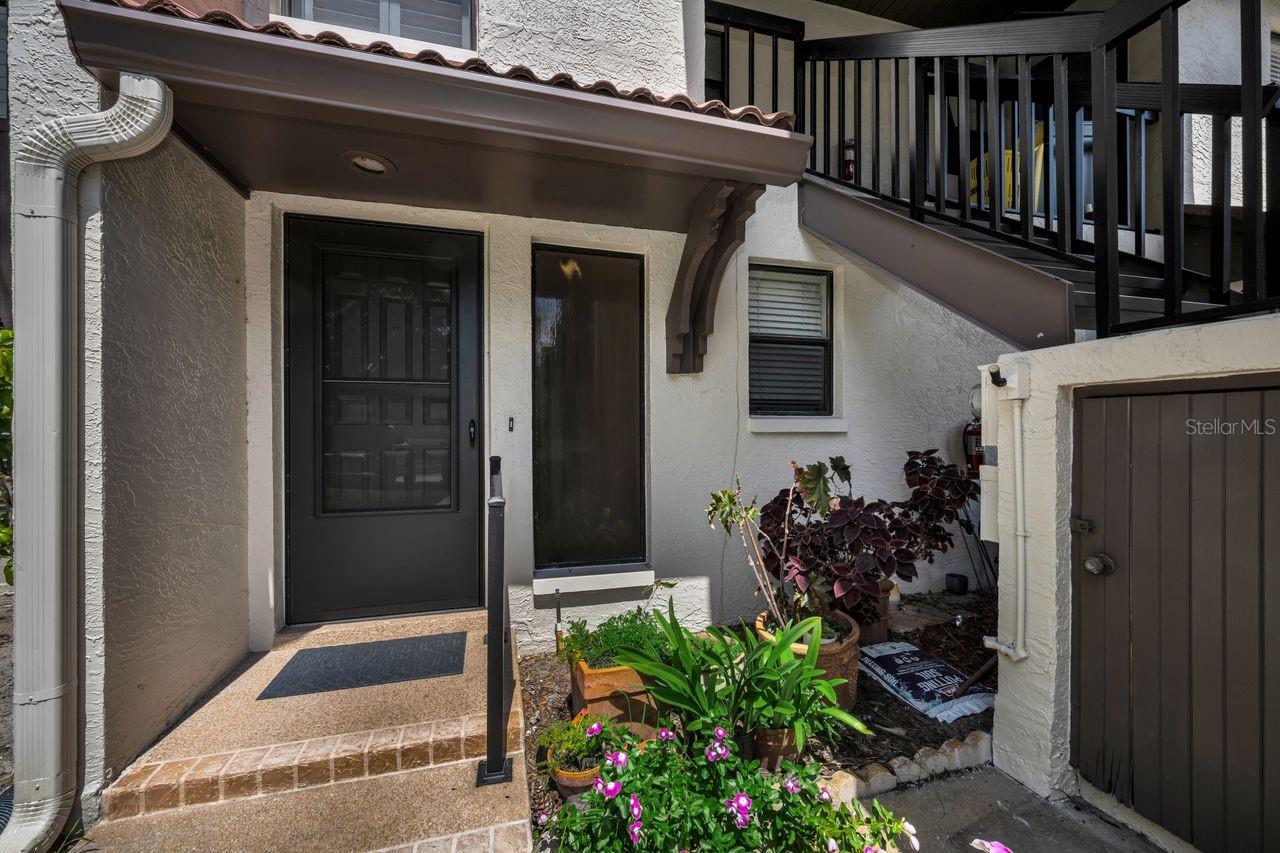 Condo entrance with a dark front door, covered walkway, and garden of flowering plants.