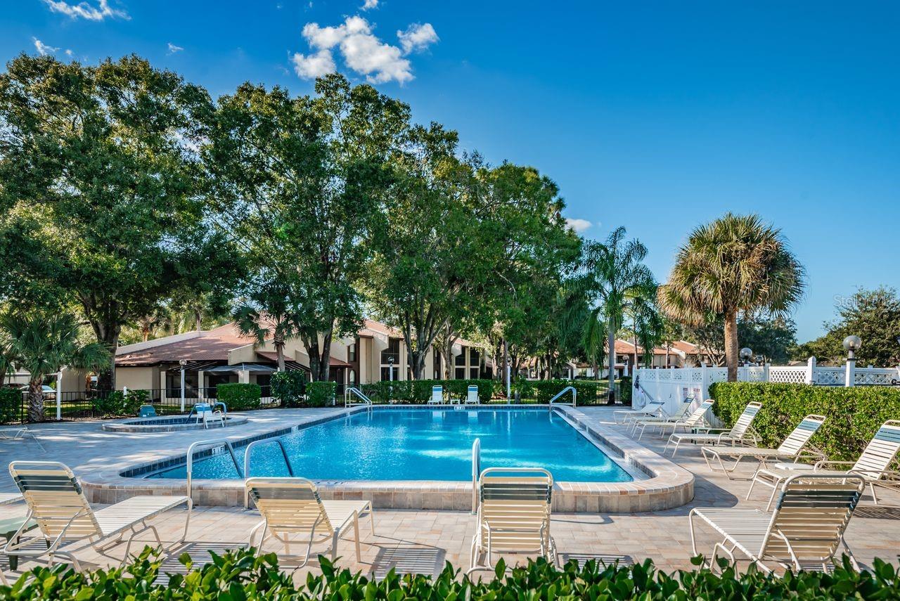 Outdoor community pool with lounge chairs, paved deck, and residences behind mature trees.