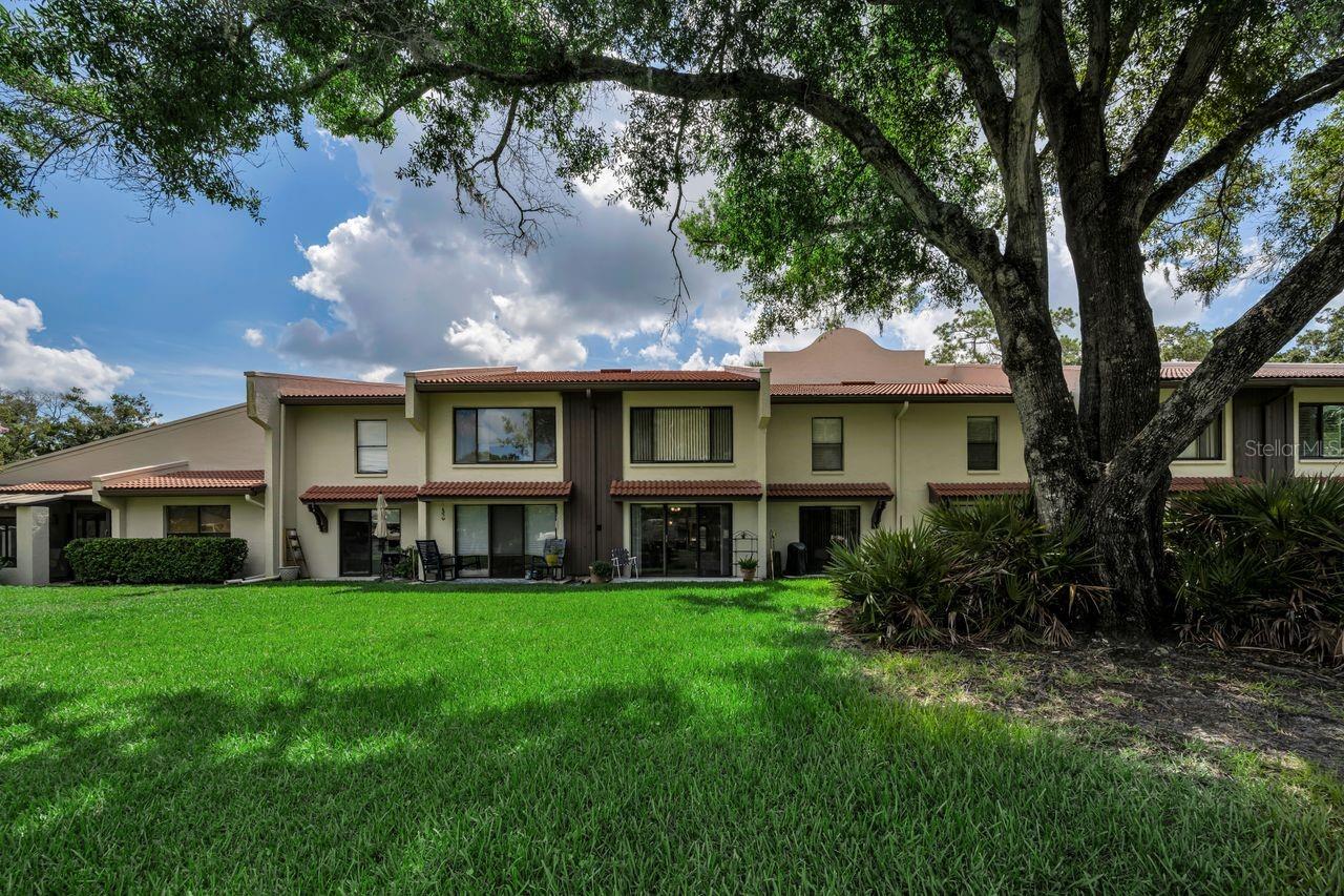 Two-story attached homes with red tile awnings, patio seating, and a lawn shaded by a large oak.