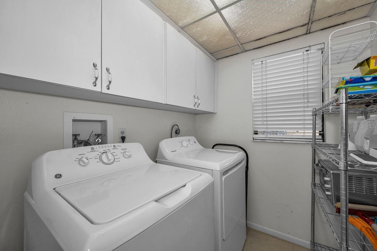 Laundry room with washer and dryer beneath overhead cabinets, beside a window and storage shelves.