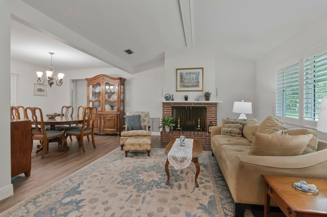 Living and dining room with a brick fireplace, beige sectional, and plantation-shuttered windows.