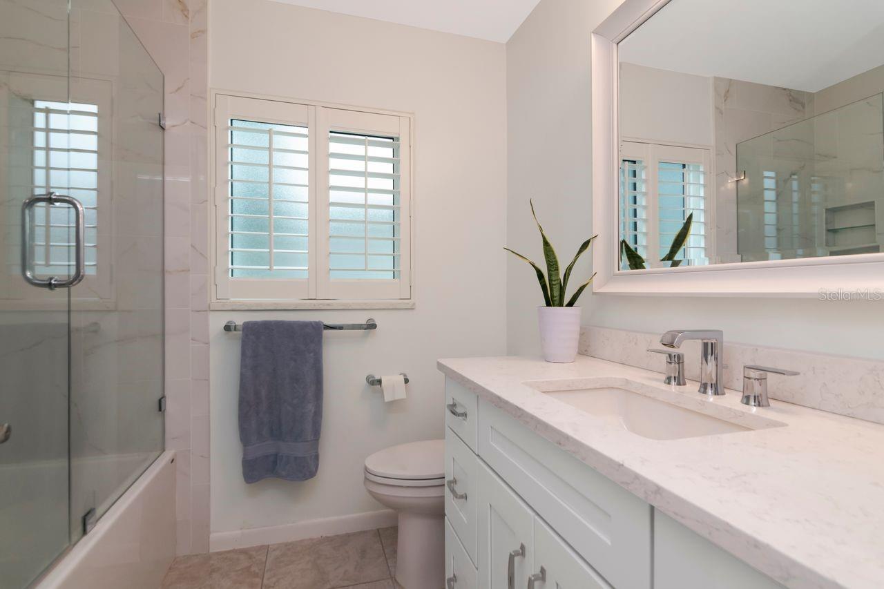 Bathroom with a glass shower, white vanity, shuttered window, and potted plant.