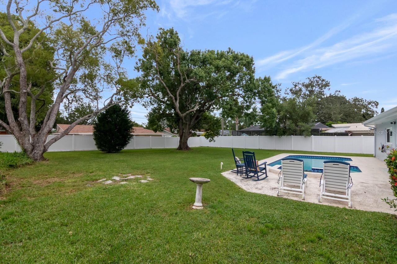 Backyard with a swimming pool, patio seating, broad lawn, mature trees, and white privacy fence.