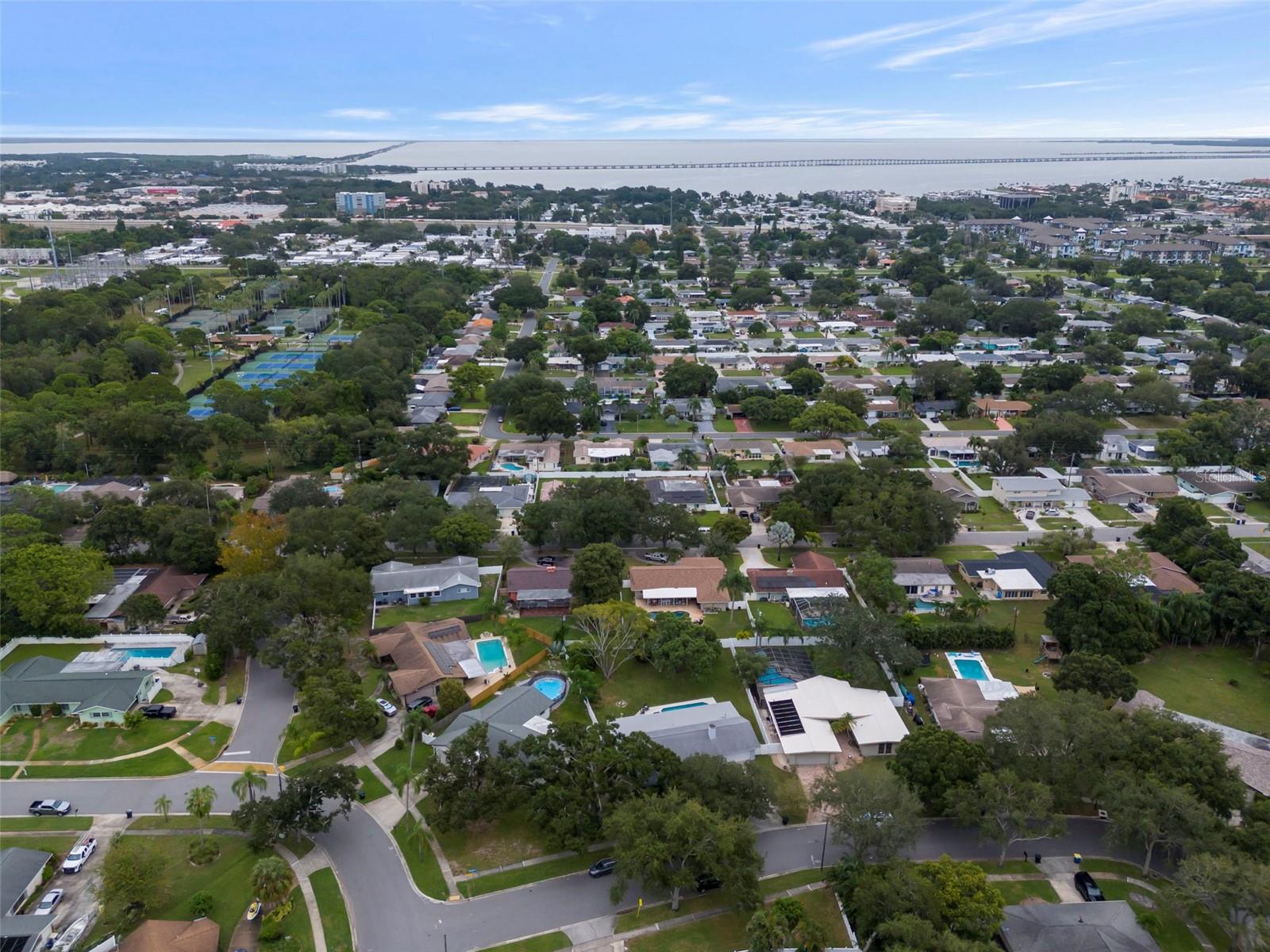 Aerial view of a Clearwater neighborhood with homes, pools, tennis courts, and a bay crossed by a bridge.