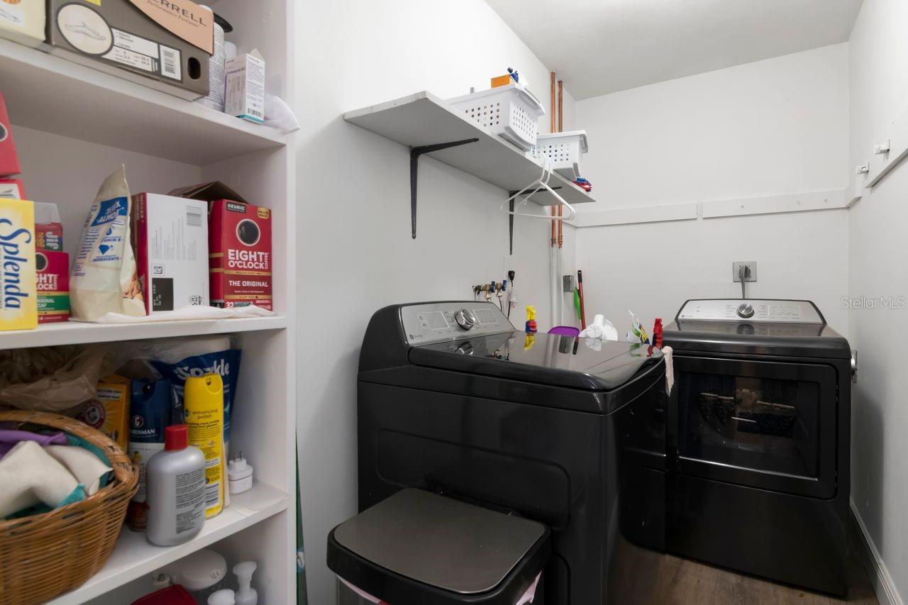 Laundry room with black washer and dryer beside shelves stocked with household supplies.