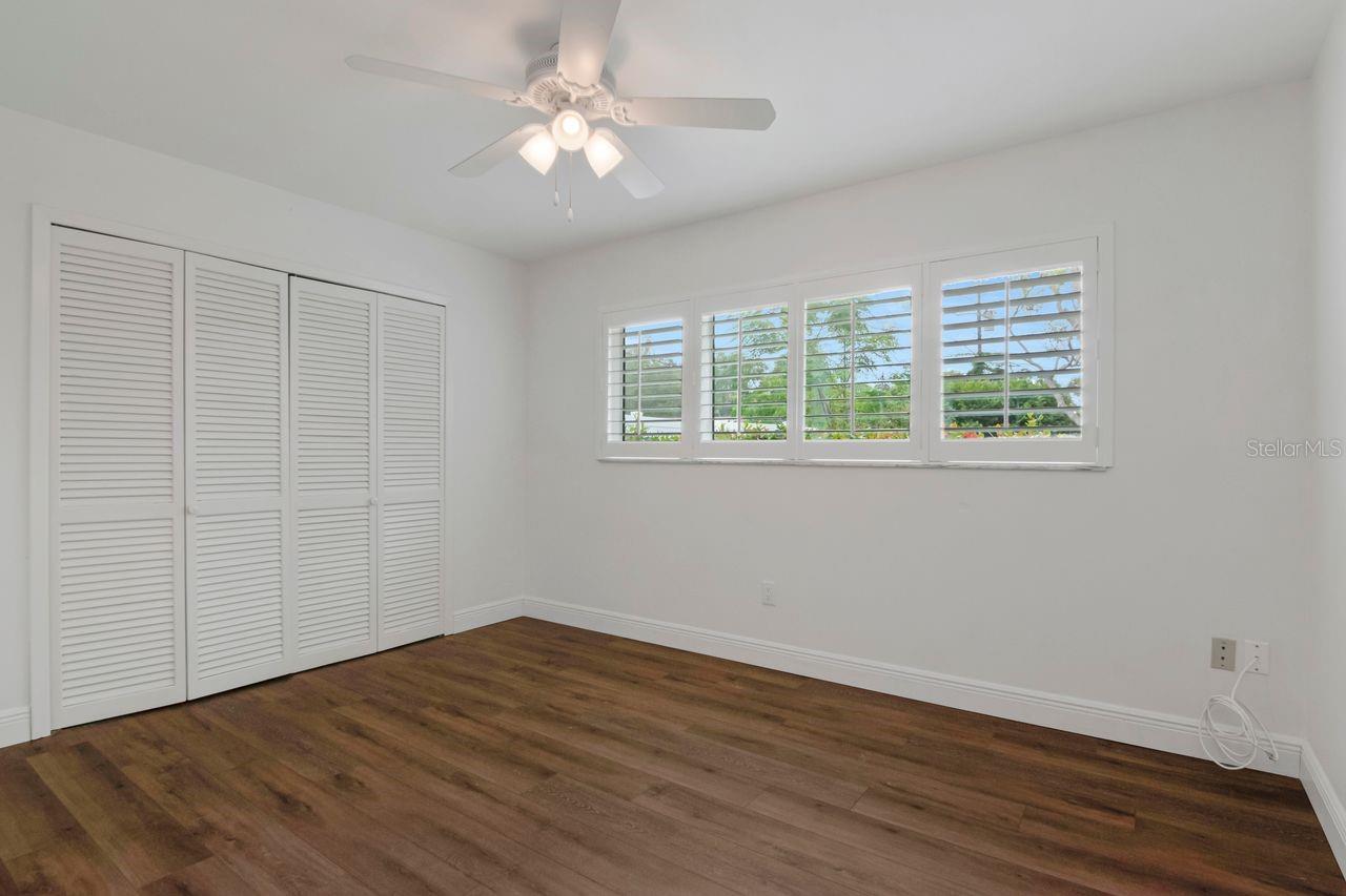 Empty bedroom with wood floors, louvered closet doors, and wide plantation-shuttered windows.