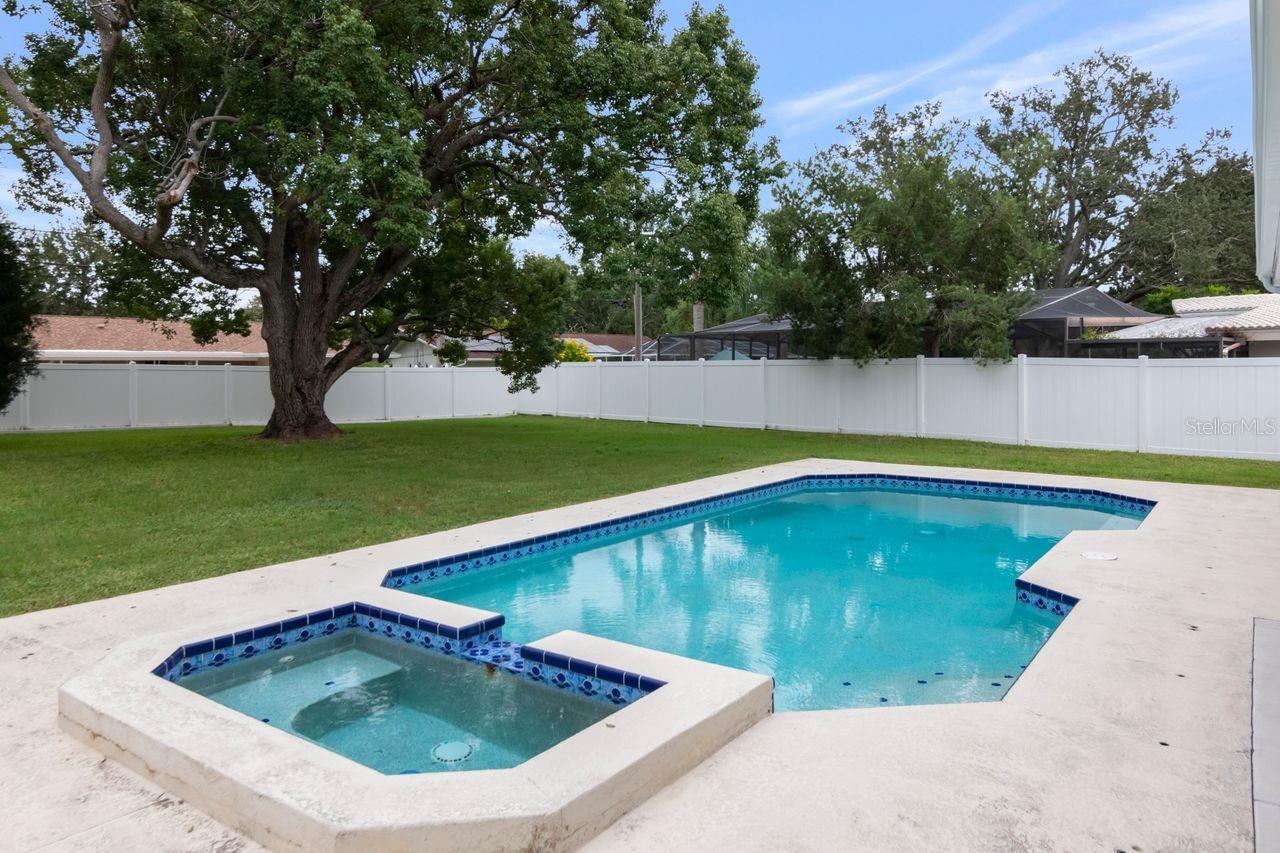 Backyard pool and raised spa beside a lawn, mature tree, and white privacy fence.