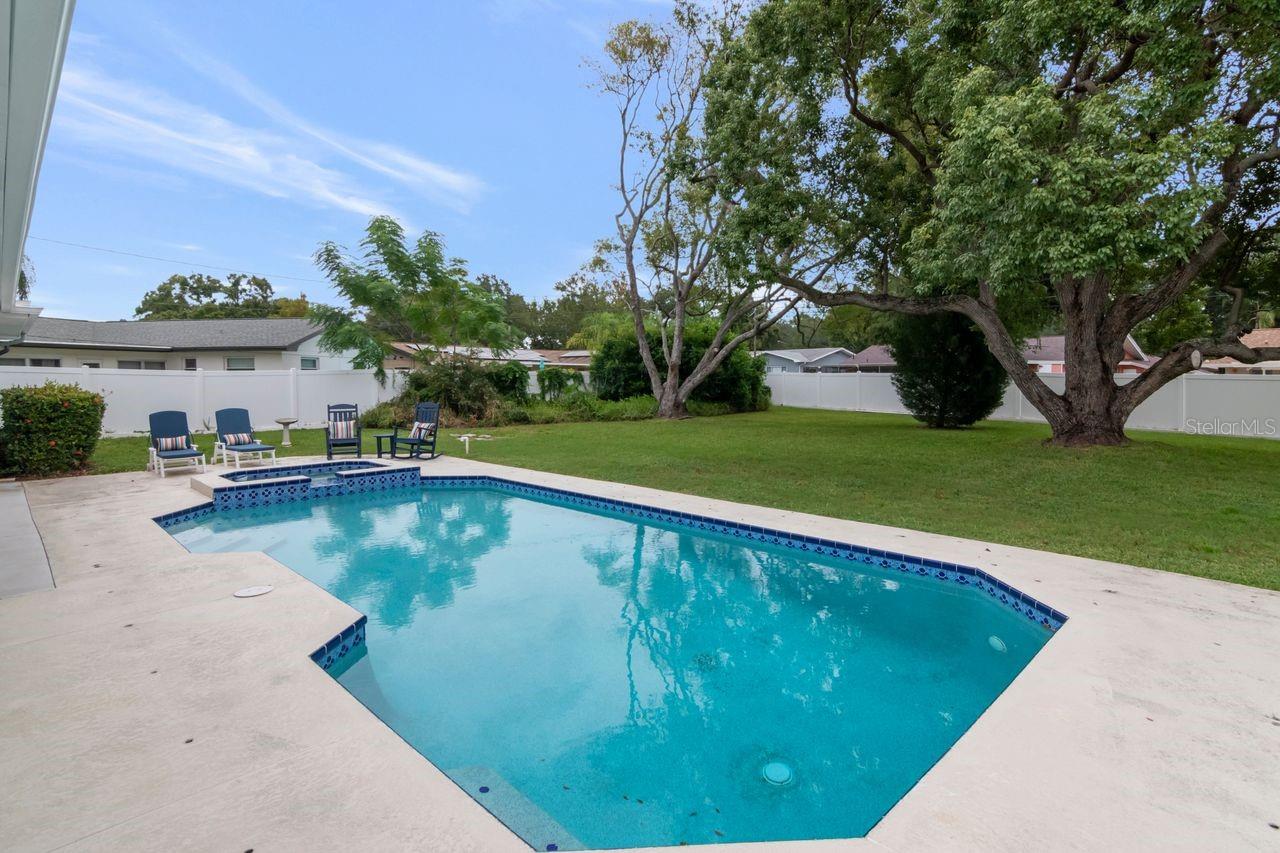 Backyard swimming pool with blue tile trim, a broad concrete deck, mature trees, and outdoor chairs.