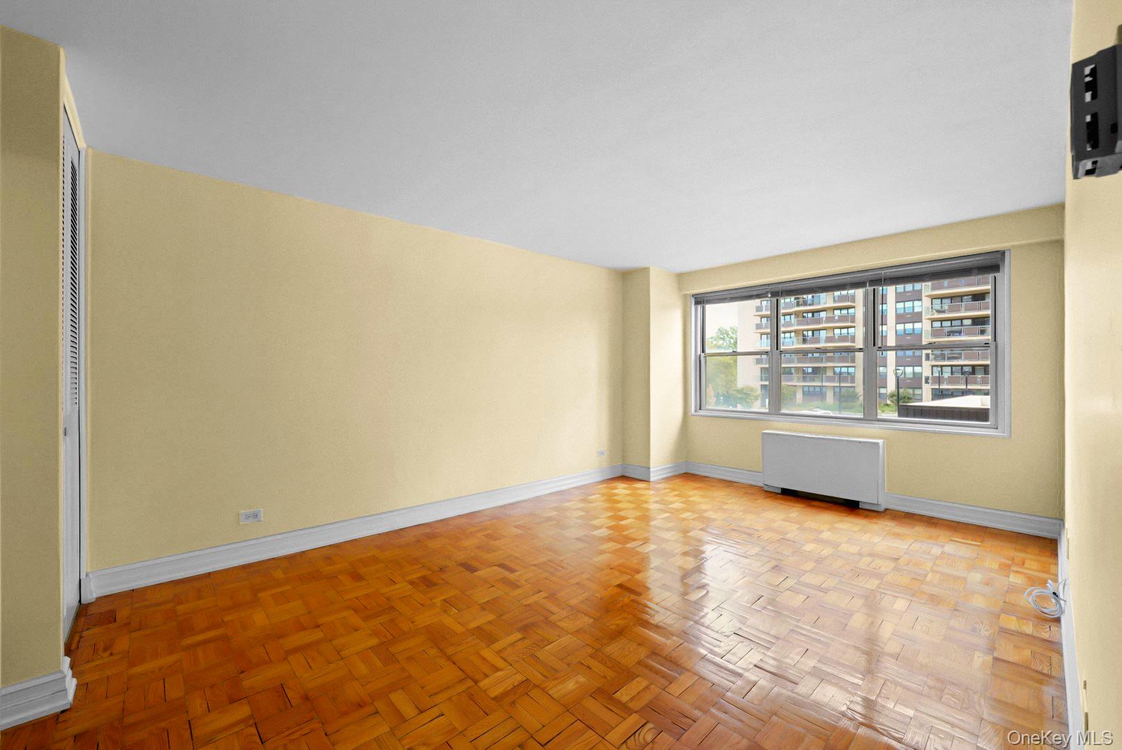 Empty living room with parquet floors, pale yellow walls, and a wide window overlooking nearby apartment buildings.