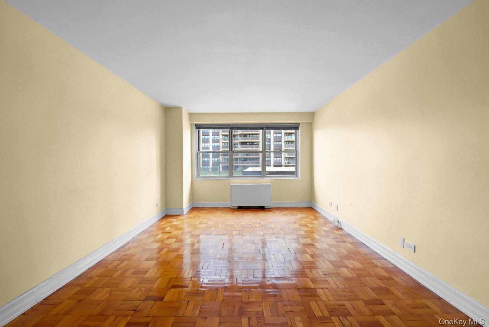 Empty living room with polished parquet floors, pale yellow walls, and a wide window above a radiator.
