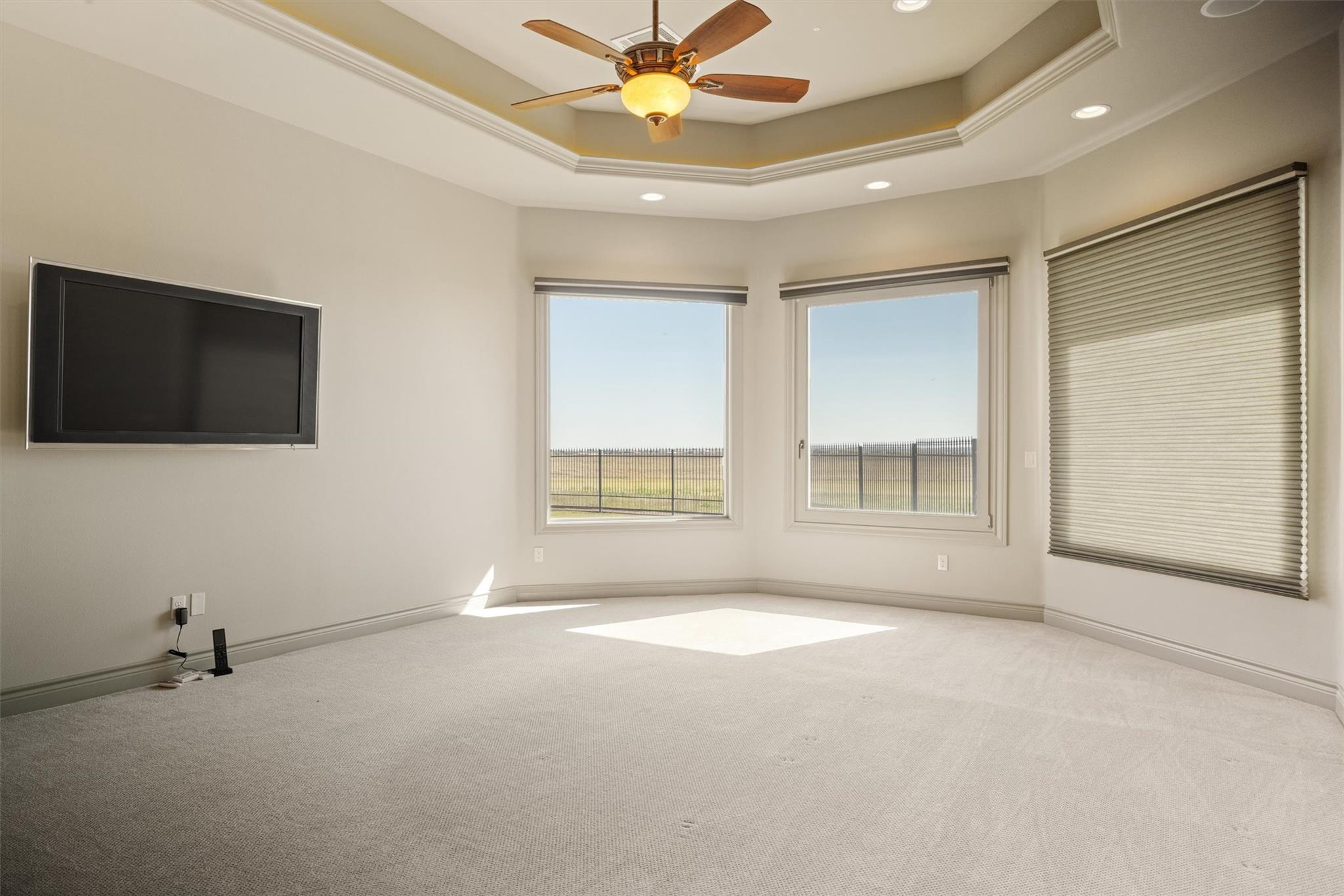 Mostly empty carpeted room with a wall-mounted television, tray ceiling, and windows overlooking a fenced area.