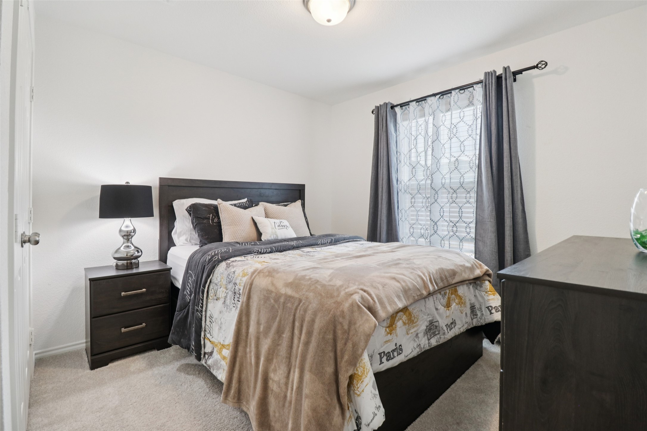 Staged bedroom with a dark wood bed, layered neutral bedding, and gray curtains beside a large window.