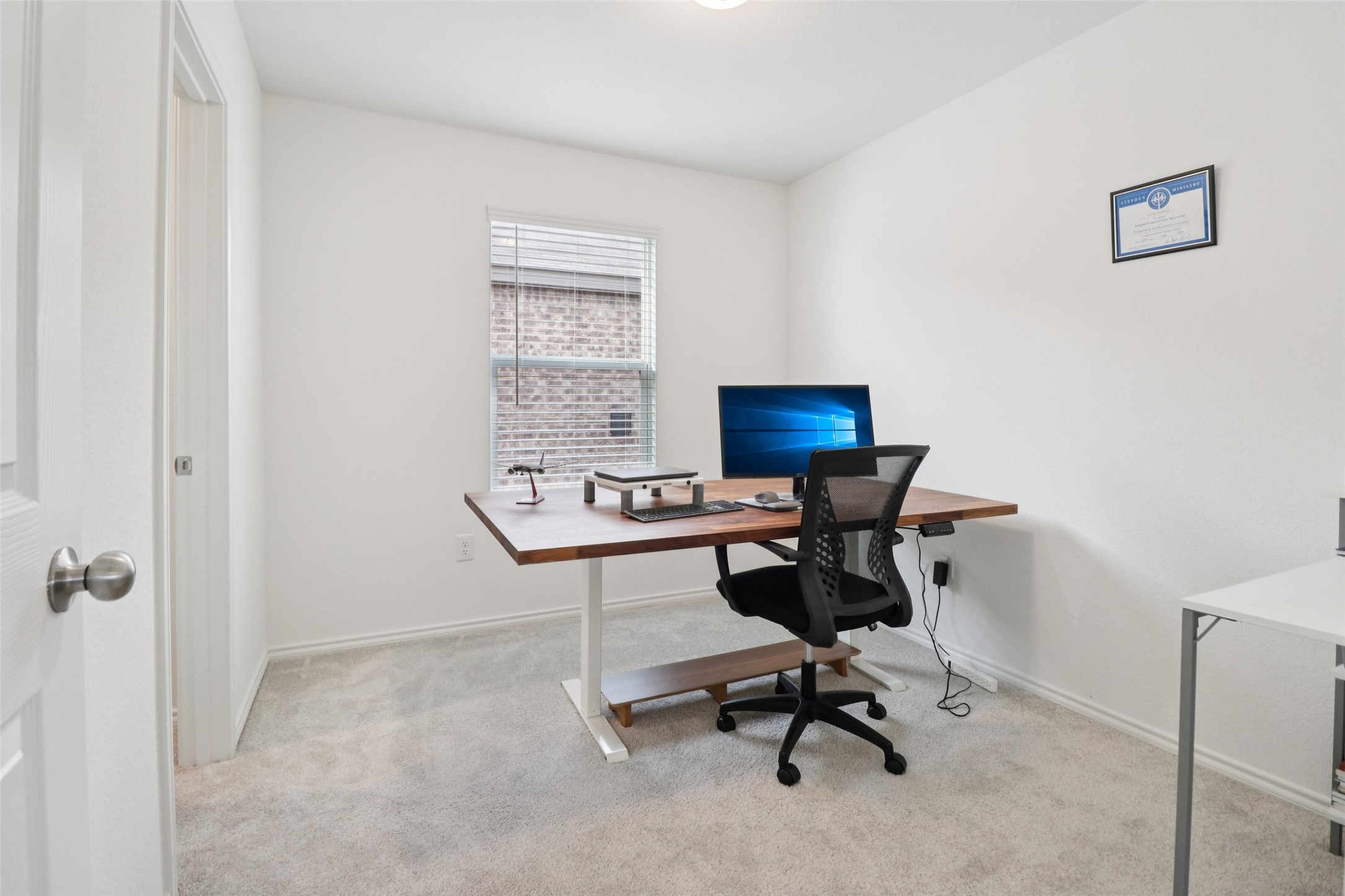 Home office with a wooden sit-stand desk, computer, and black mesh chair beside a window.