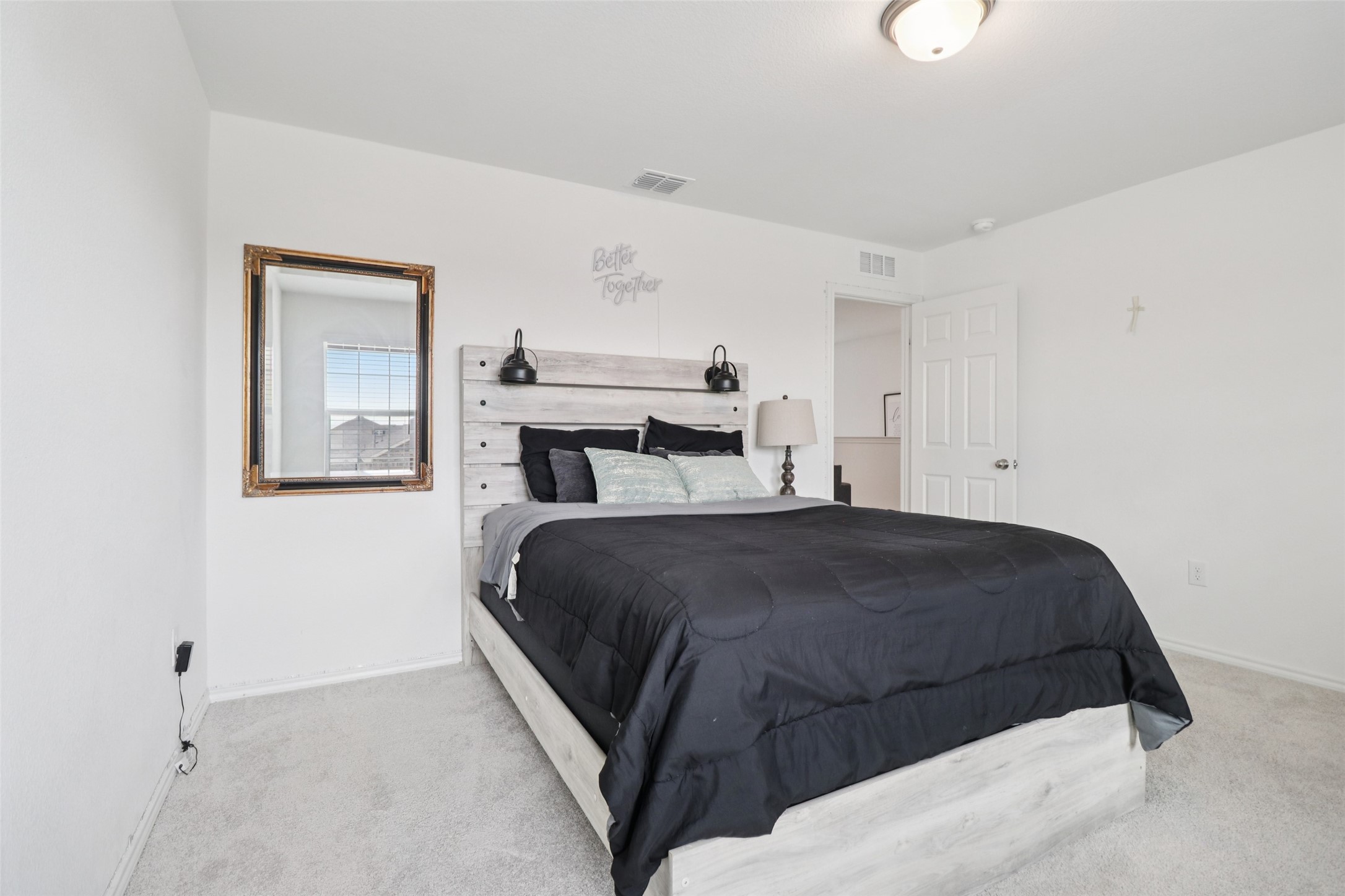 Furnished bedroom with a large bed, light wood frame, dark quilt, and framed wall mirror.