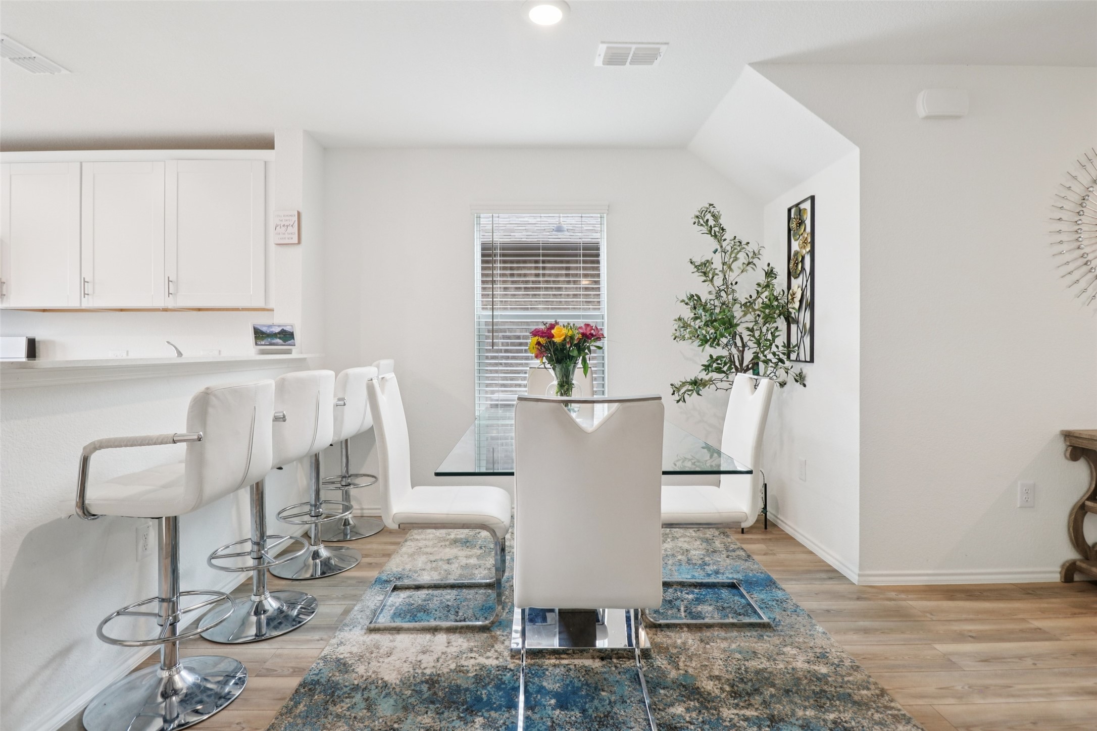 Dining area with a glass-top table, white chairs, blue rug, and kitchen counter with bar stools.