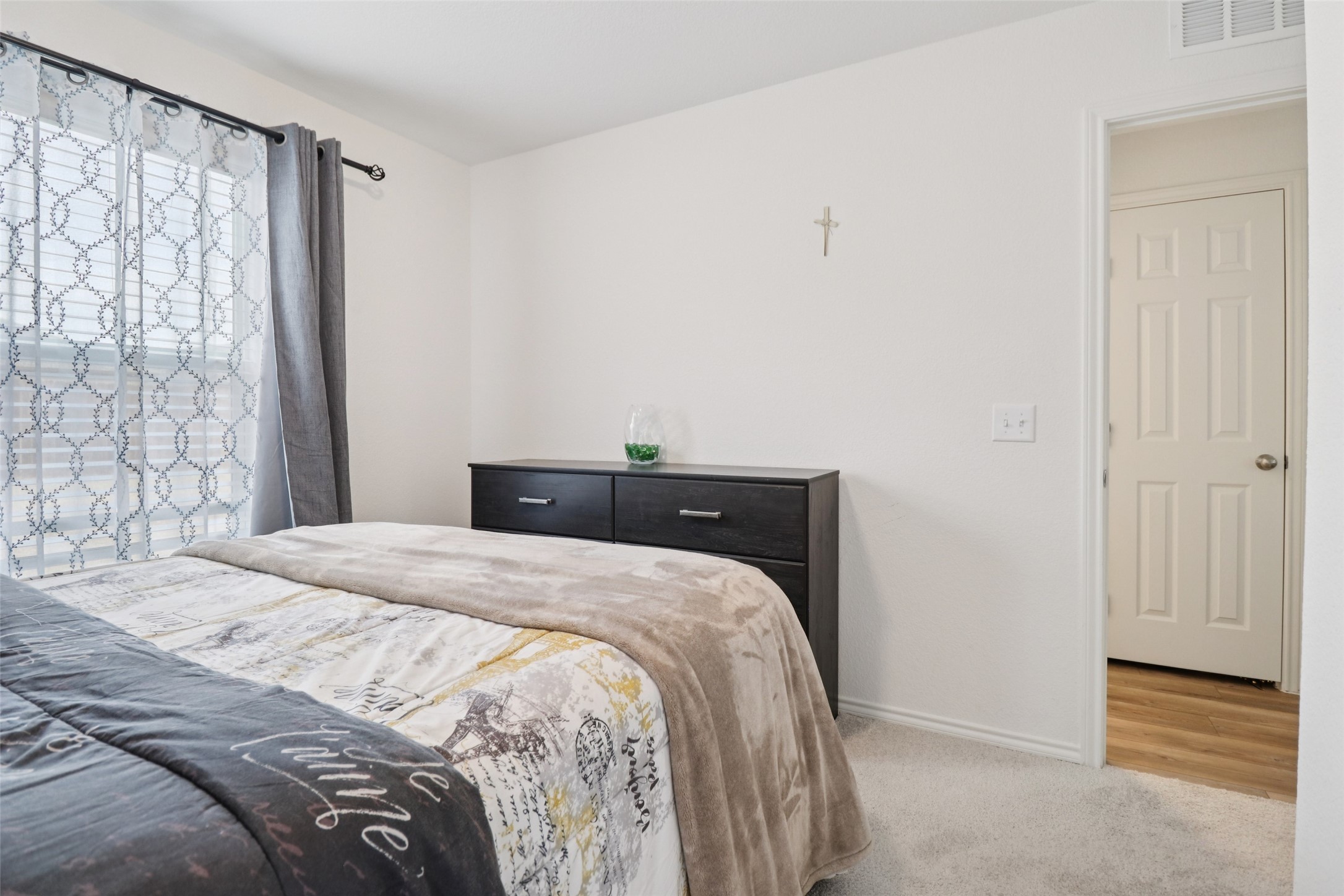 Furnished bedroom with patterned bedding, a dark dresser, and a window with layered curtains.
