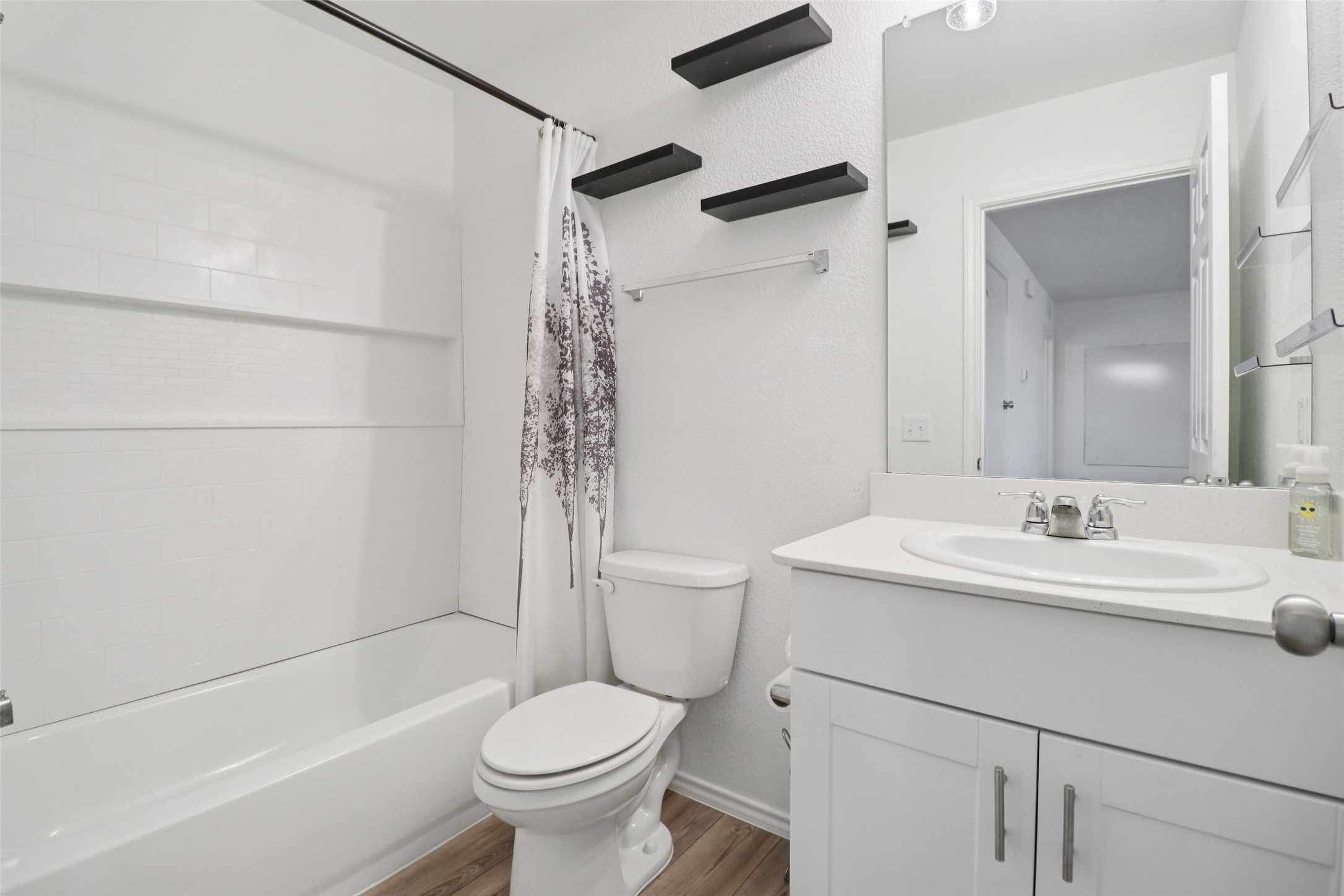 White bathroom with a tiled bathtub, patterned shower curtain, toilet, and vanity with a large mirror.