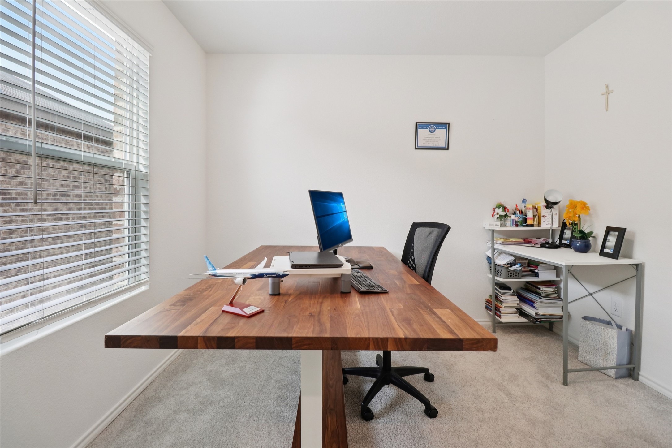Home office with a large wooden desk, computer setup, model airplane, and window with white blinds.
