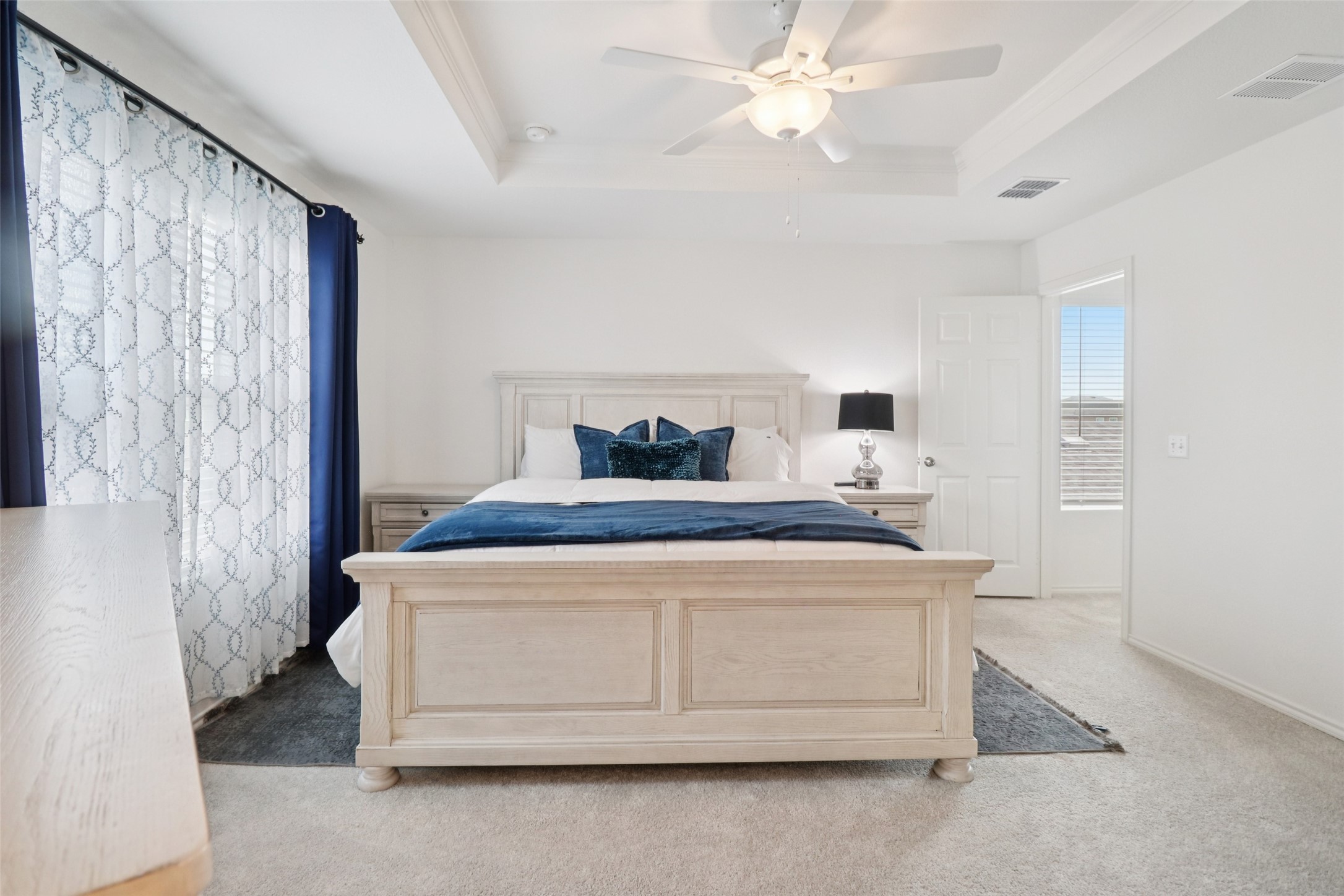 Staged bedroom with a light wood bed, navy bedding, patterned curtains, and a ceiling fan.