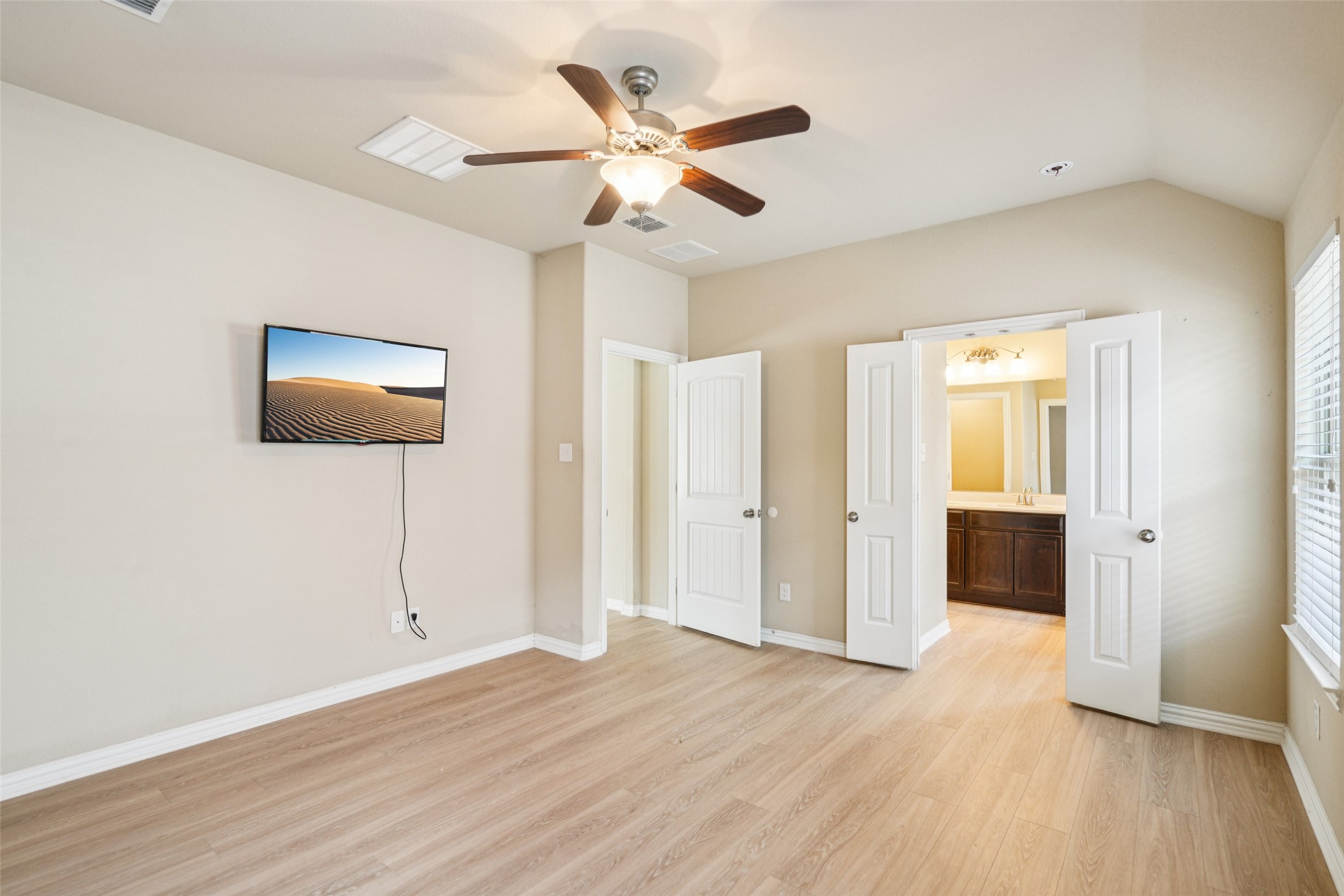 Mostly empty bedroom with a ceiling fan, wall-mounted TV, wood flooring, and open doors to an adjoining bathroom.