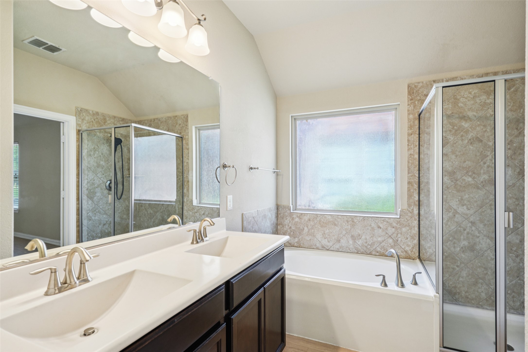 Bathroom with a dark double vanity, tub beneath a frosted window, and tiled glass shower.