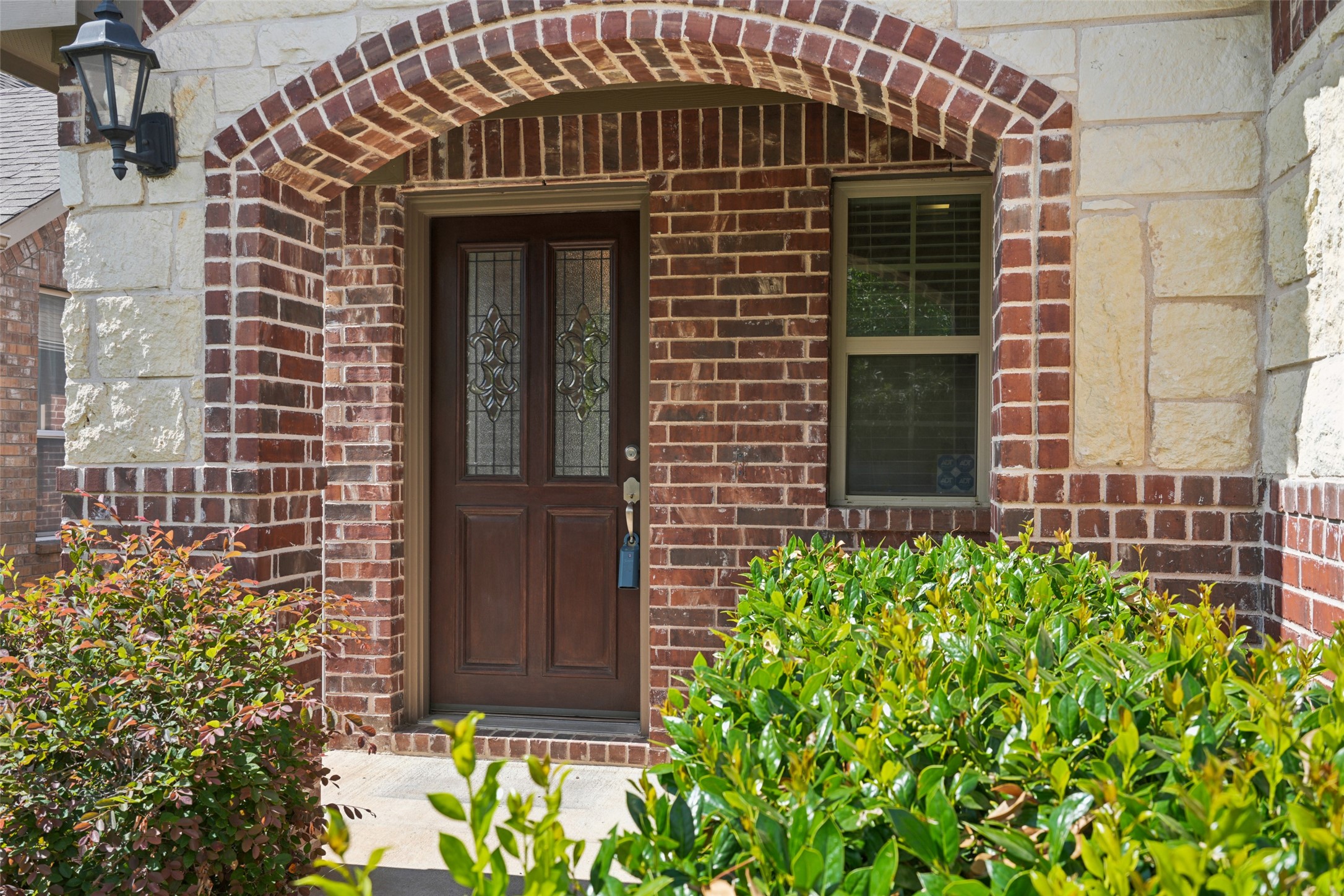 Front entrance with a decorative double door beneath a brick arch, framed by stonework and shrubs.
