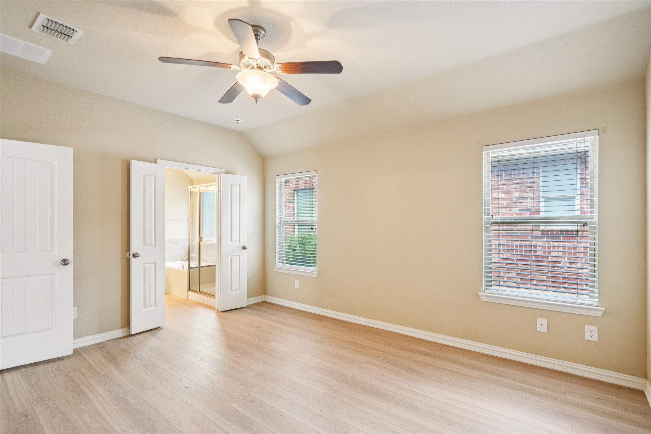 Empty bedroom with light wood floors, a ceiling fan, and open doors to an adjoining bathroom.