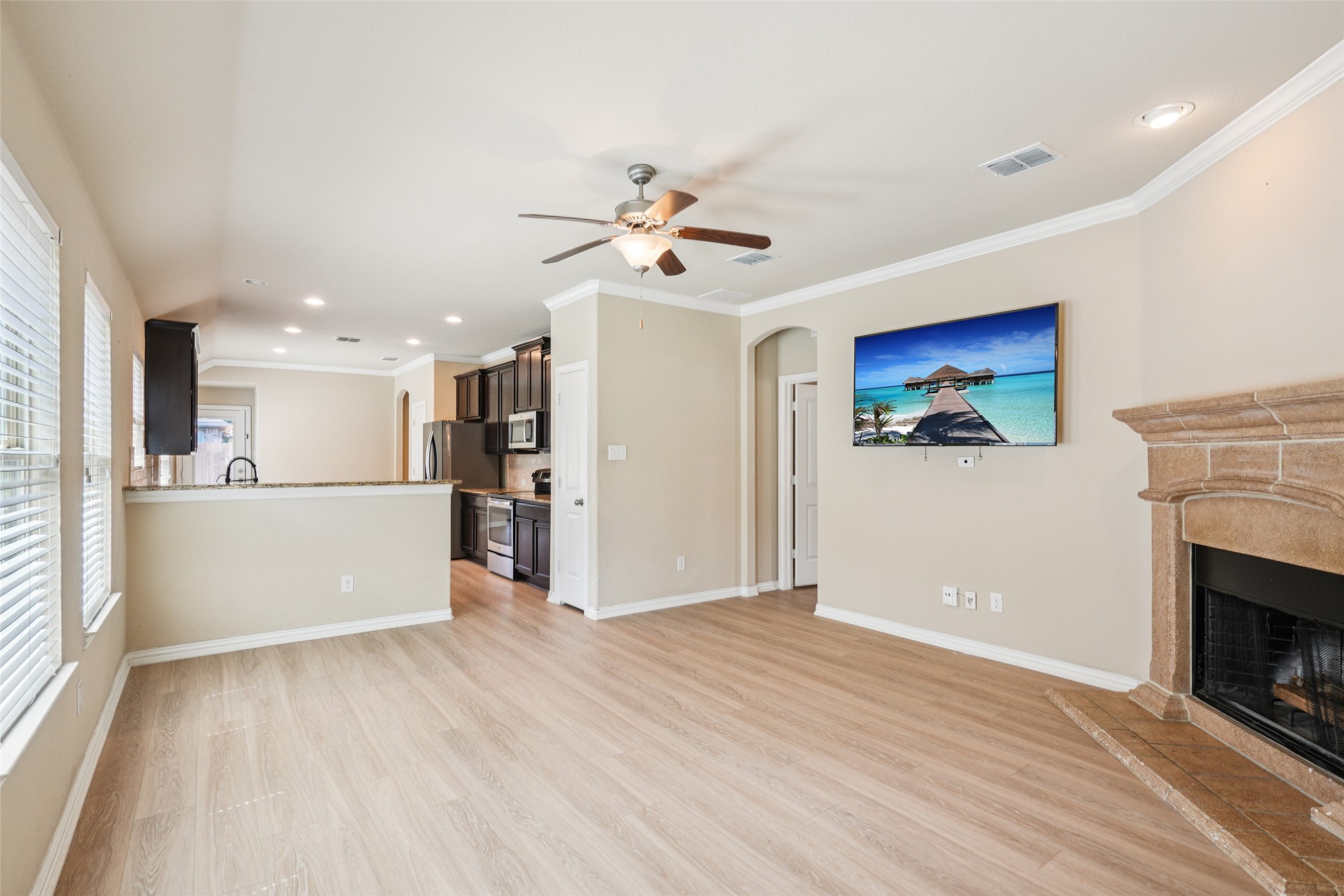 Living room with a stone fireplace and light wood flooring, open to a kitchen with dark cabinets.