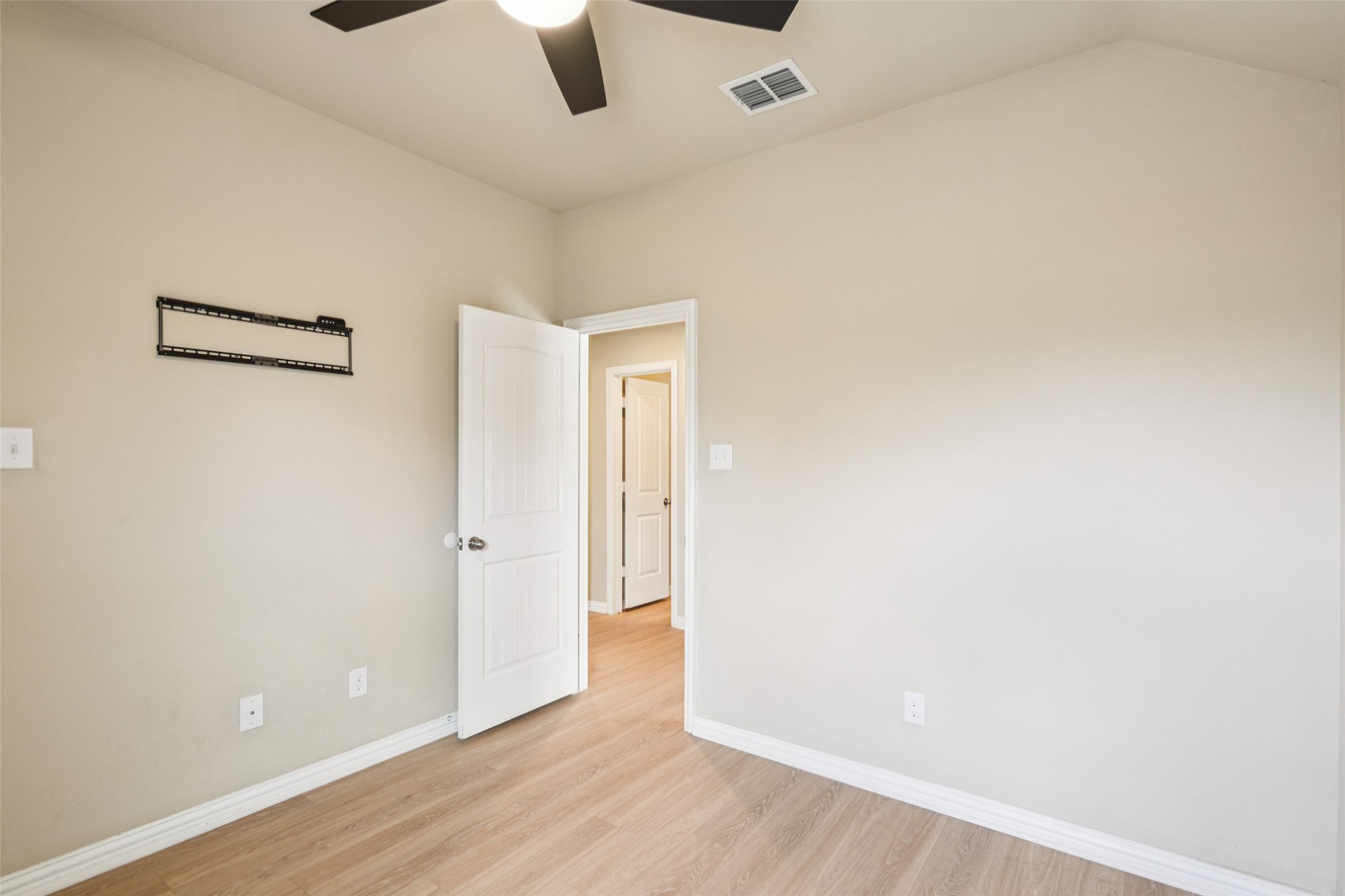 Empty bedroom with light wood flooring, ceiling fan, and wall-mounted TV bracket.