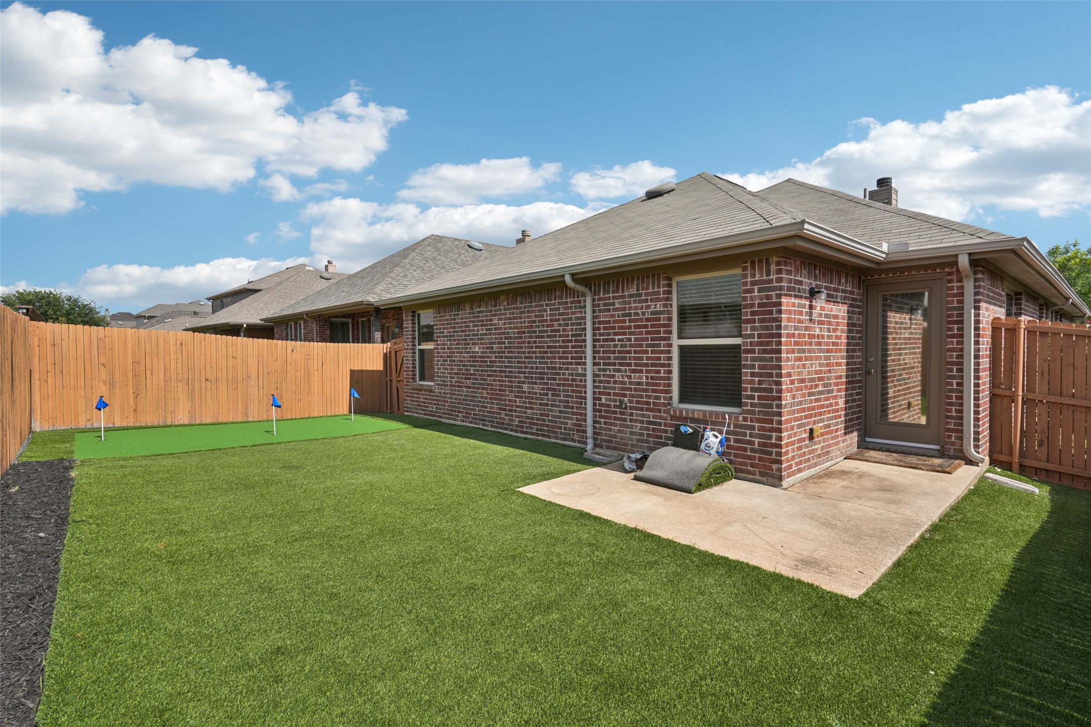 Red-brick home's fenced backyard with a large lawn, small putting green, and concrete patio.