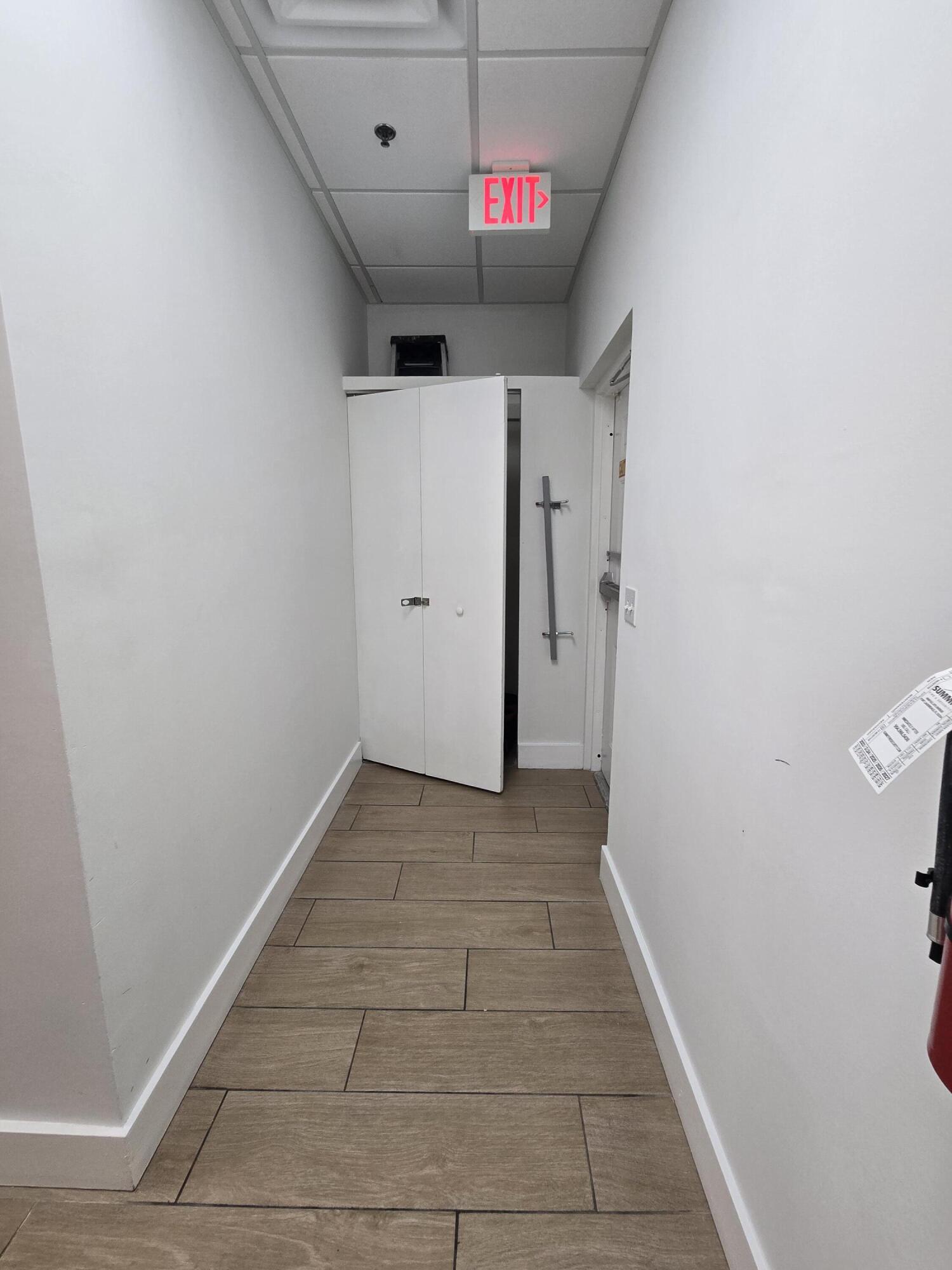 Empty hallway with brown tile flooring, a partially open door, and an illuminated EXIT sign.