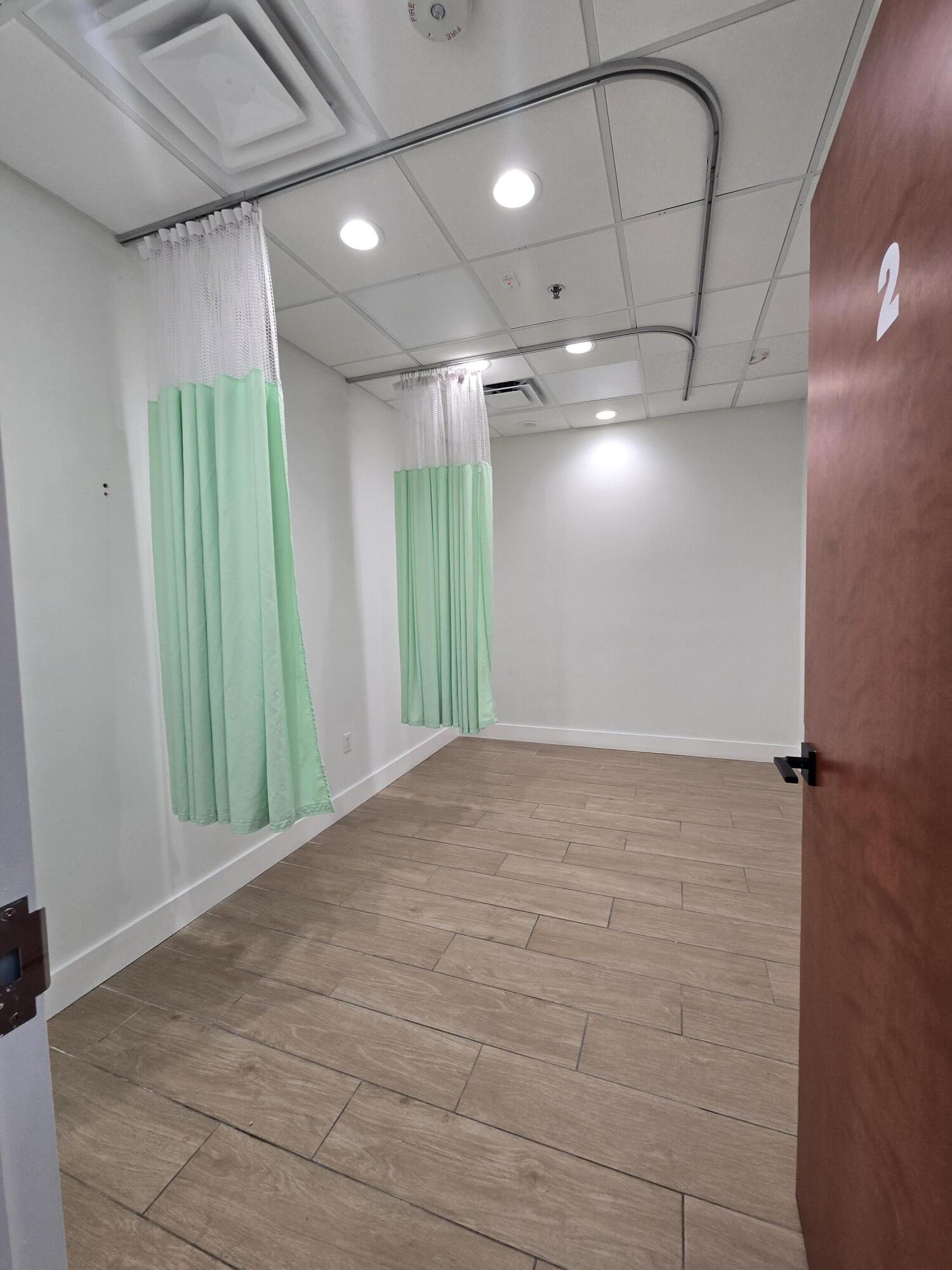 Medical examination room with mint-green privacy curtains and wood-look tile flooring.