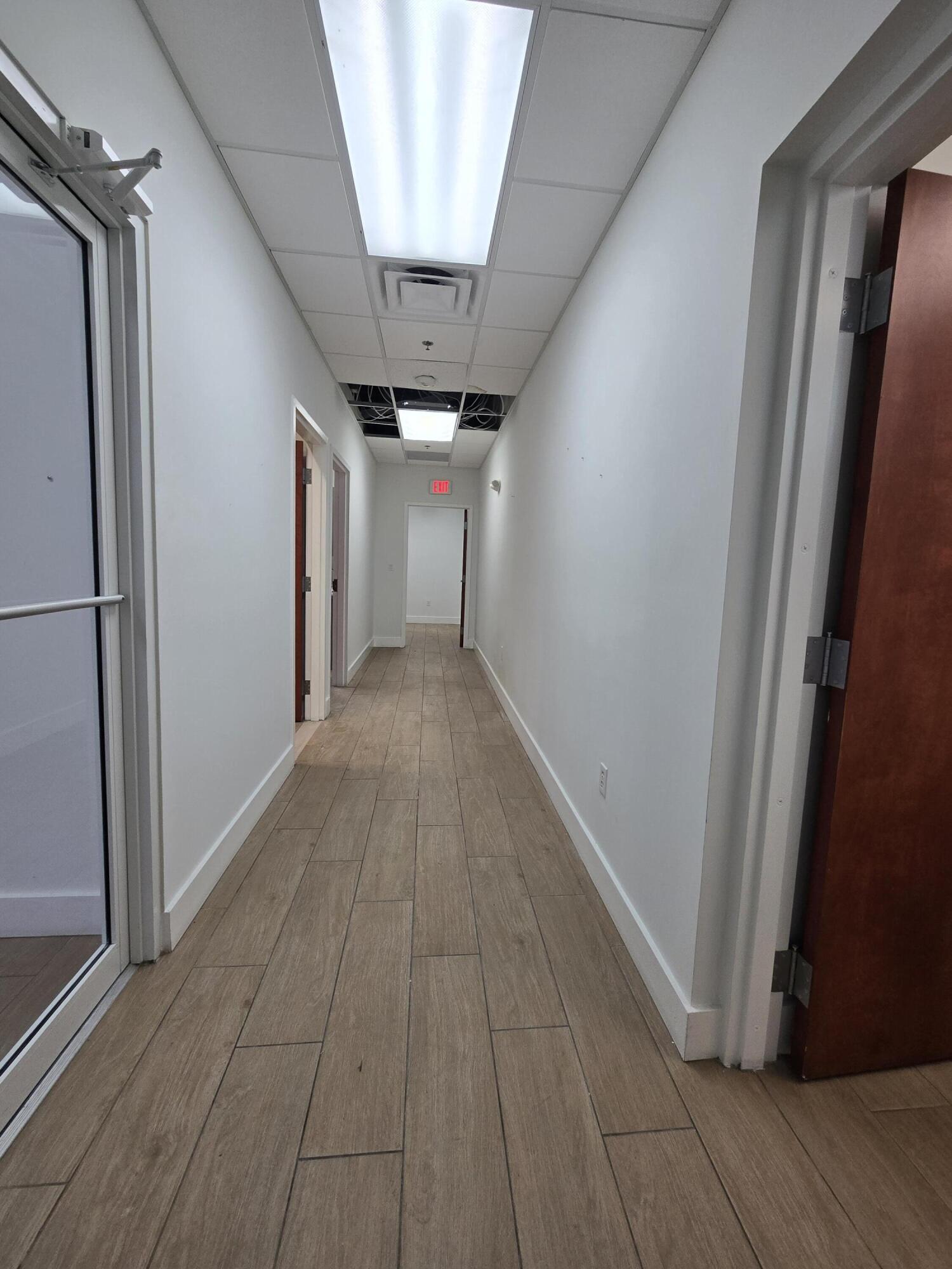Empty commercial hallway with wood-look tile flooring and exposed ceiling wiring.