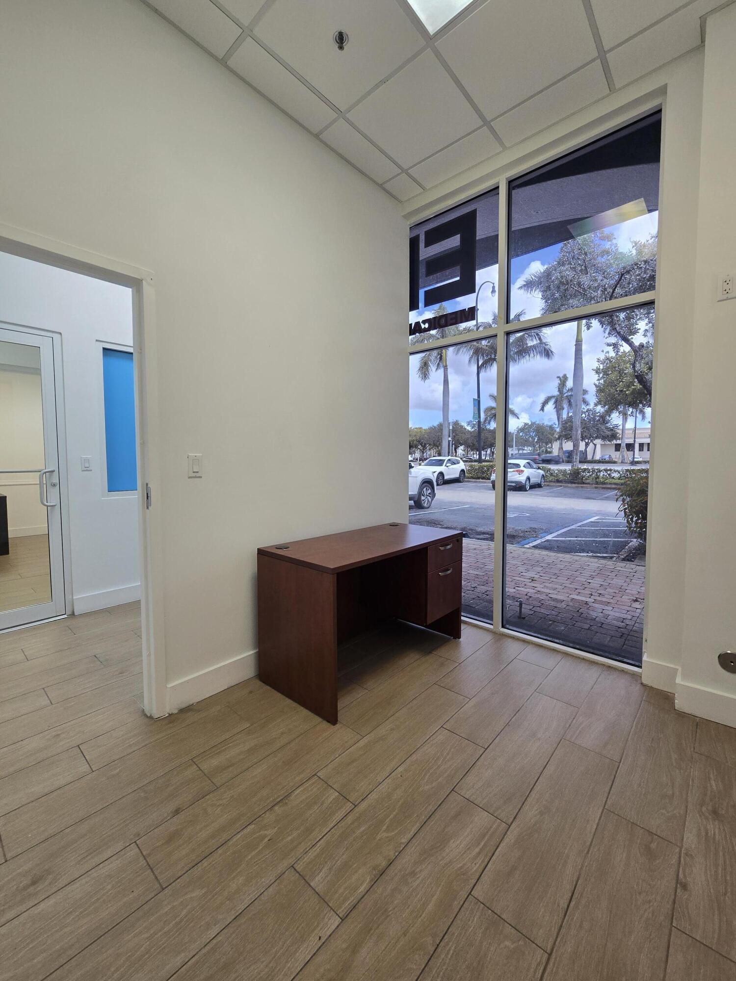 Small office with a dark wood desk beside tall windows overlooking a palm-lined parking lot.