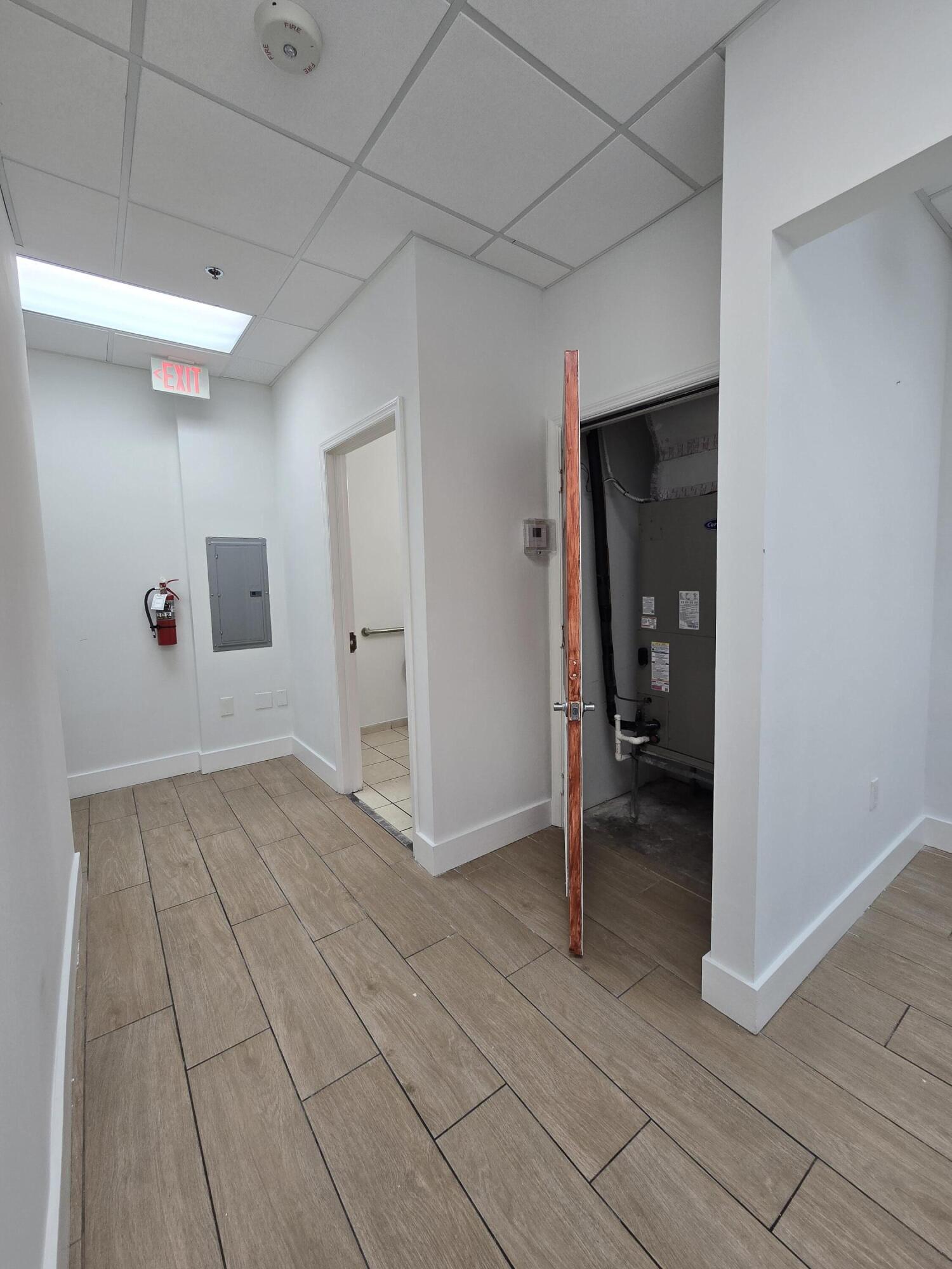 Commercial hallway with an open HVAC closet, beige tile flooring, and an illuminated exit sign.