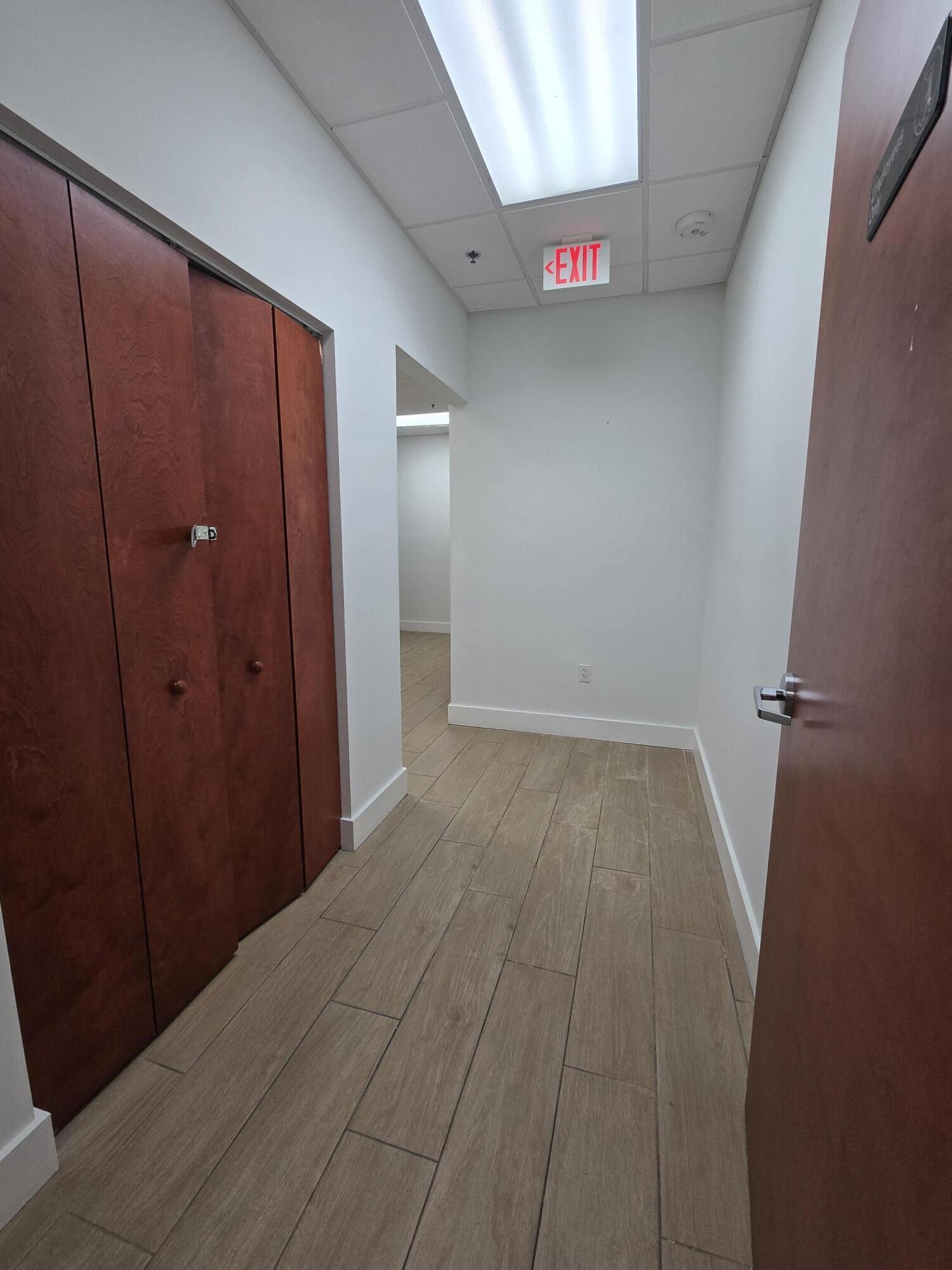 Empty hallway with wood-look tile floors, reddish-brown cabinets, and an illuminated EXIT sign.