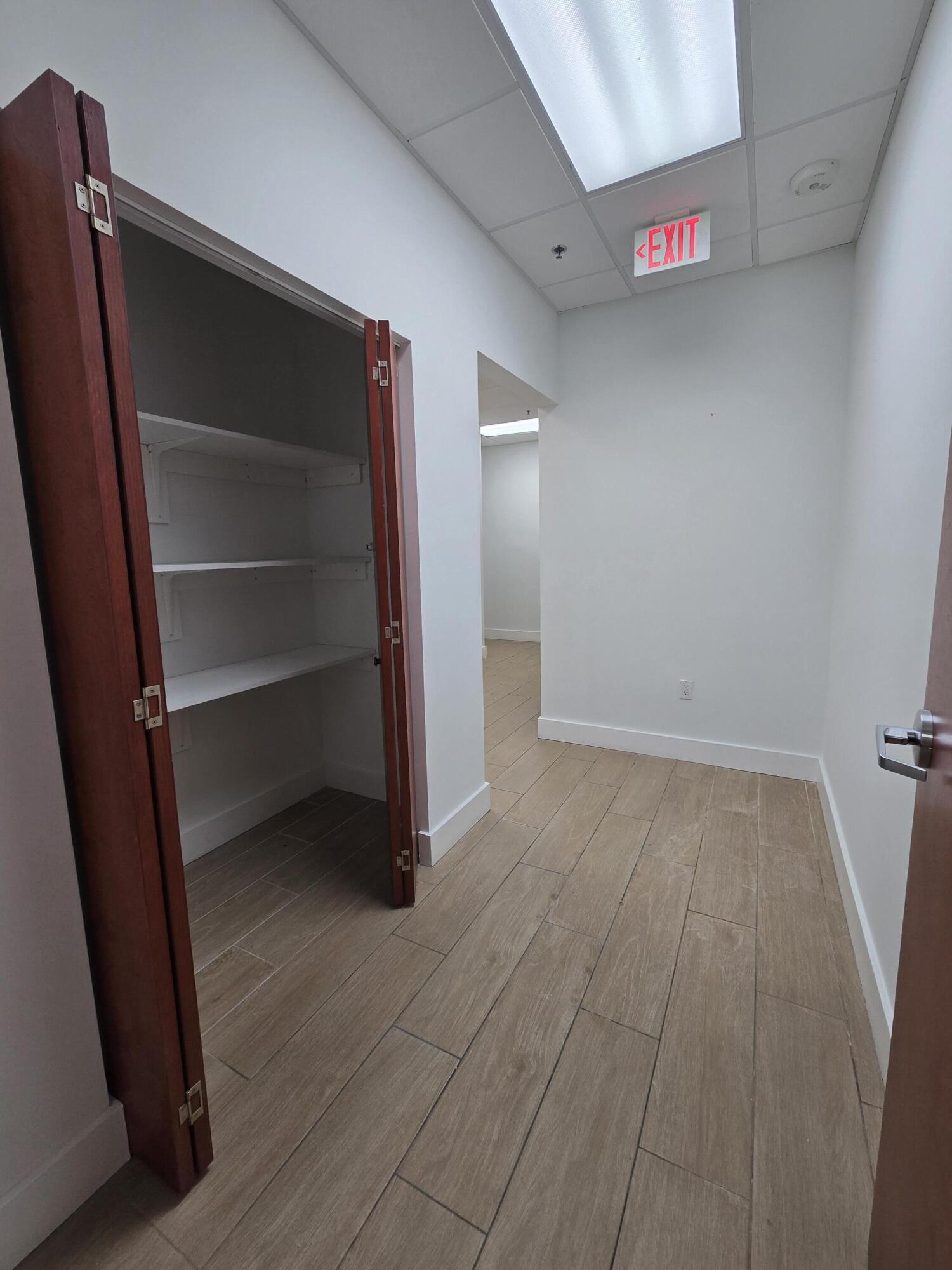 Small empty interior with an open built-in shelving cabinet, tan tile flooring, and an illuminated EXIT sign.