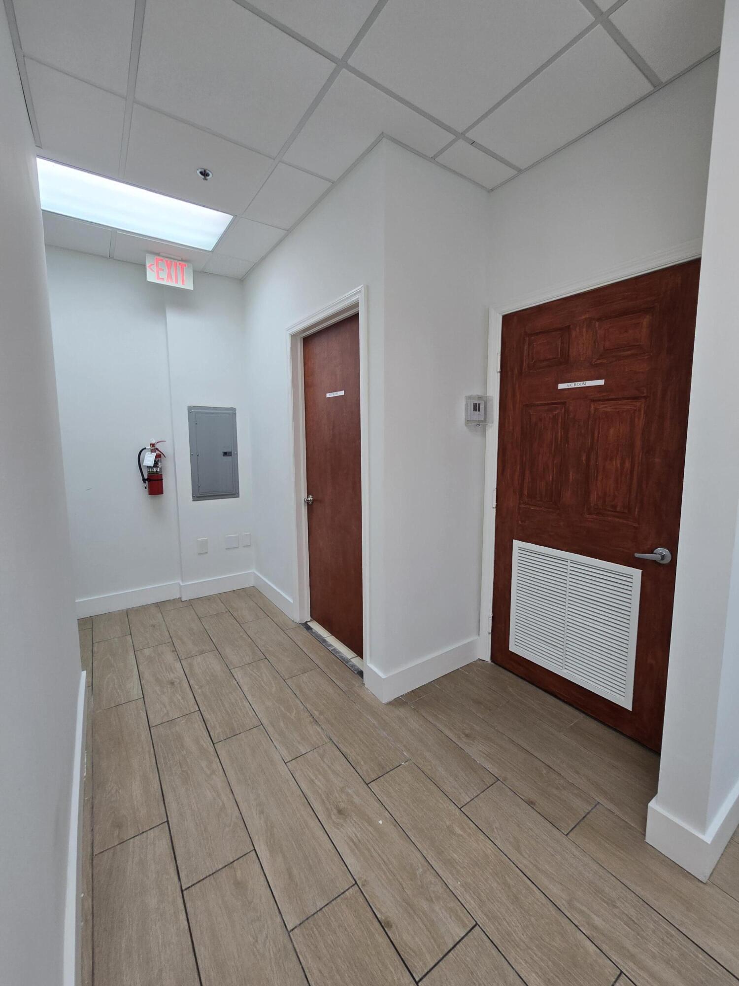 Commercial hallway with tan tile flooring, wooden doors, and an illuminated exit sign.