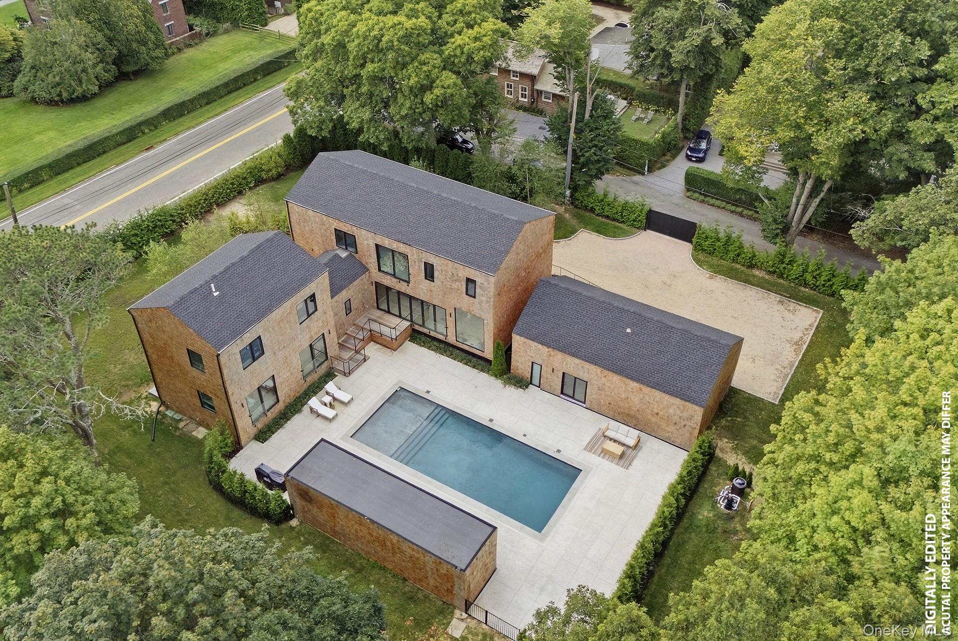 Aerial view of a modern shingle-sided home with a pool, paved patio, and gravel driveway.
