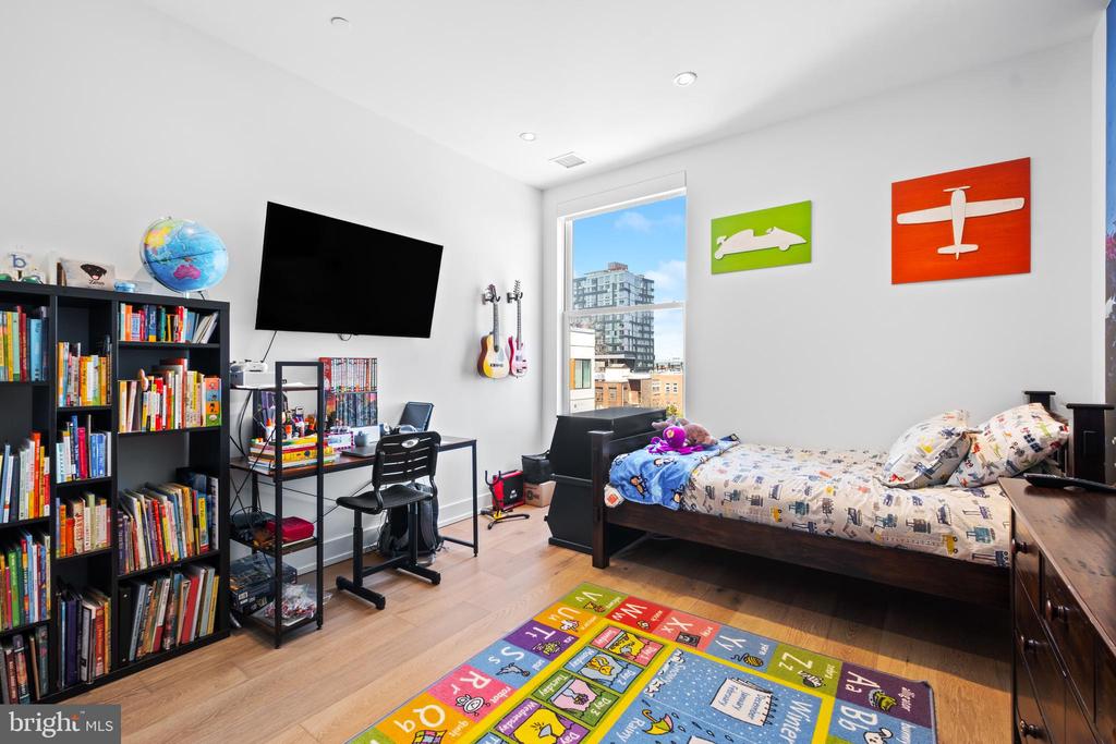 Bedroom with bookcases, a desk, guitars, and a large window overlooking city buildings.
