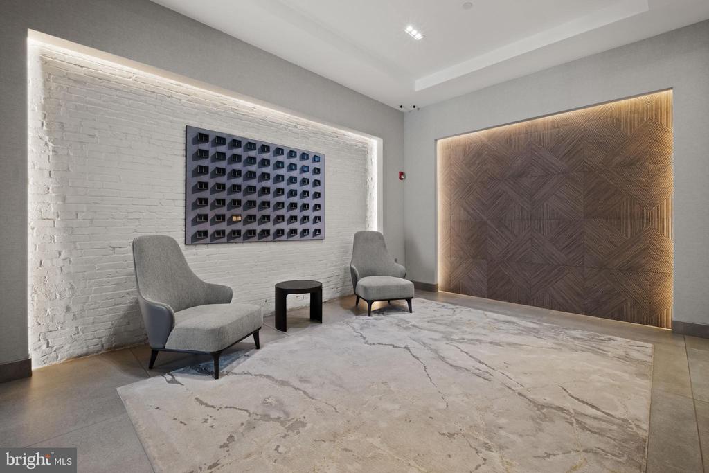 Lobby seating area with gray chairs, white brick, geometric wood paneling, and a large stone-patterned rug.