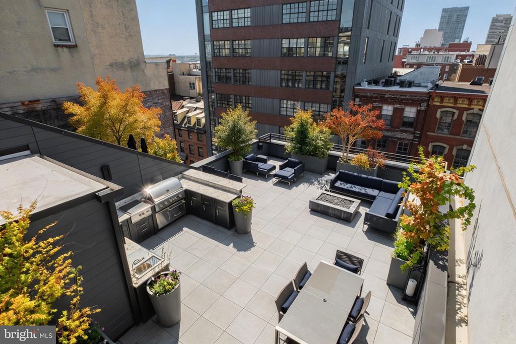 Rooftop terrace with dining and lounge seating, built-in grill, and autumn planters among city buildings.