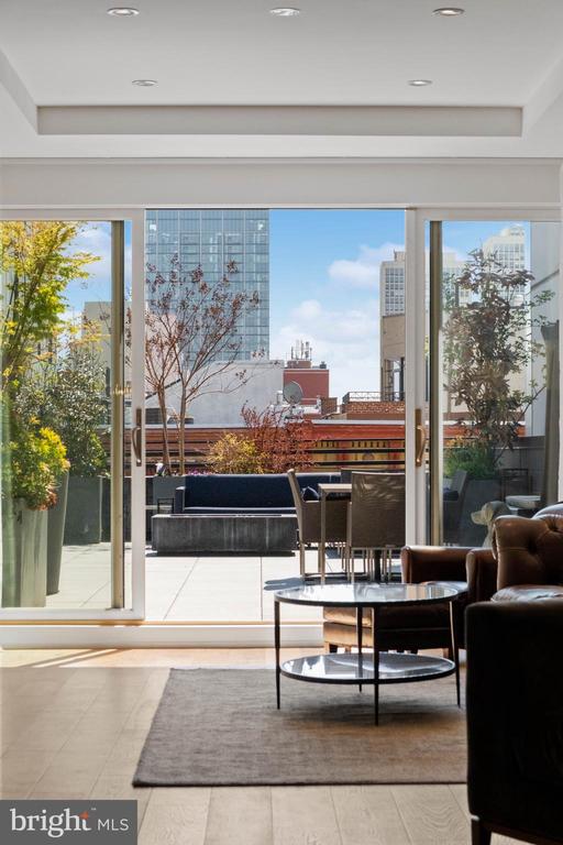 Living room with dark leather seating and glass coffee tables overlooking a furnished terrace and city buildings.
