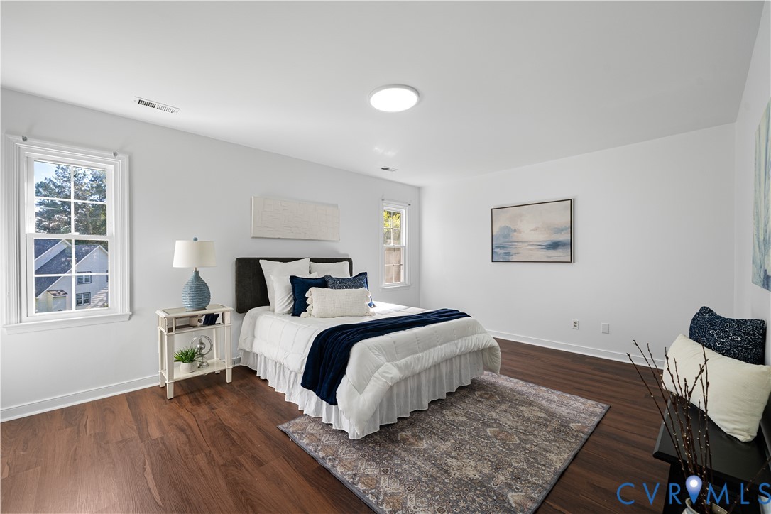 Staged bedroom with a white bed, navy accents, dark wood floors, and two windows.