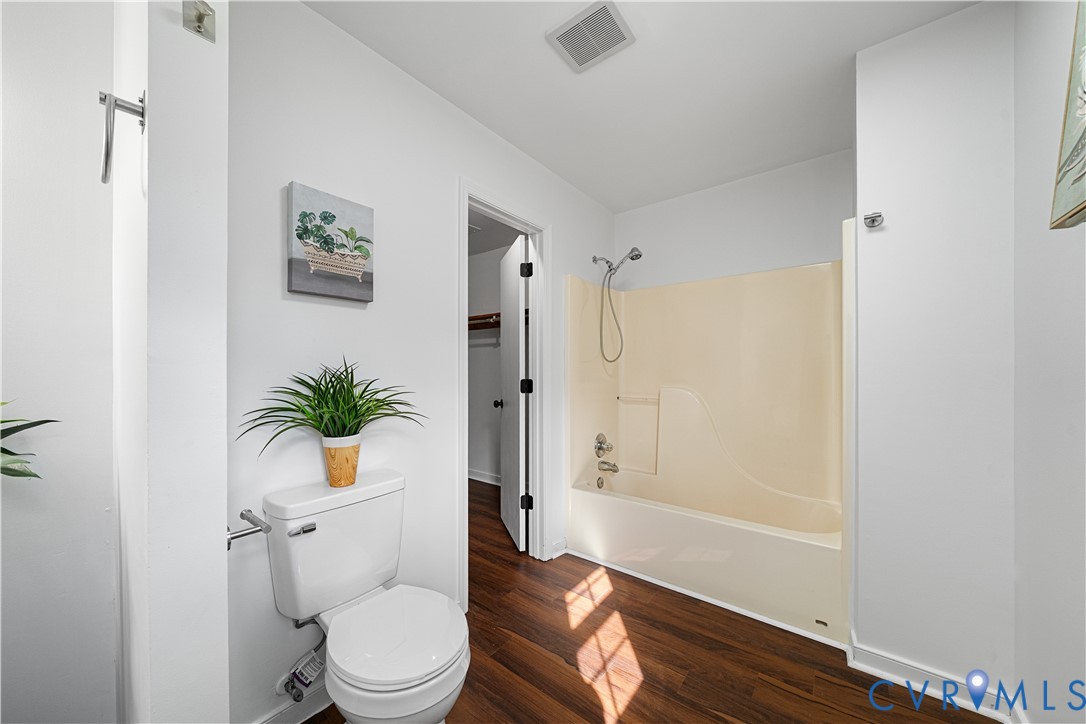 Bathroom with a cream tub and shower, white toilet, dark wood flooring, and a potted plant.