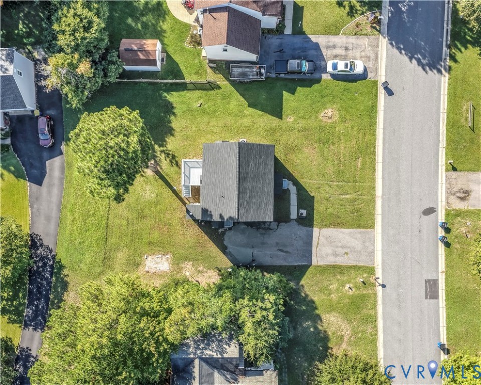 Aerial view of a house on a grassy lot beside a residential street, with mature trees and a driveway.