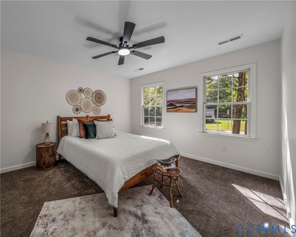 Staged bedroom with a wooden bed, white bedding, dark ceiling fan, and windows overlooking a green yard.