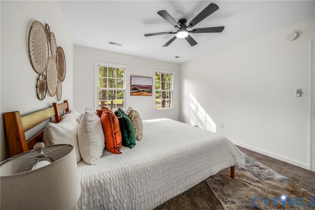 Furnished bedroom with a white bed, colorful pillows, woven wall decor, and two sunlit windows.