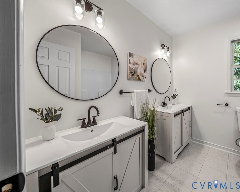Bathroom with two white vanities, oversized round mirrors, dark fixtures, and light gray tile flooring.